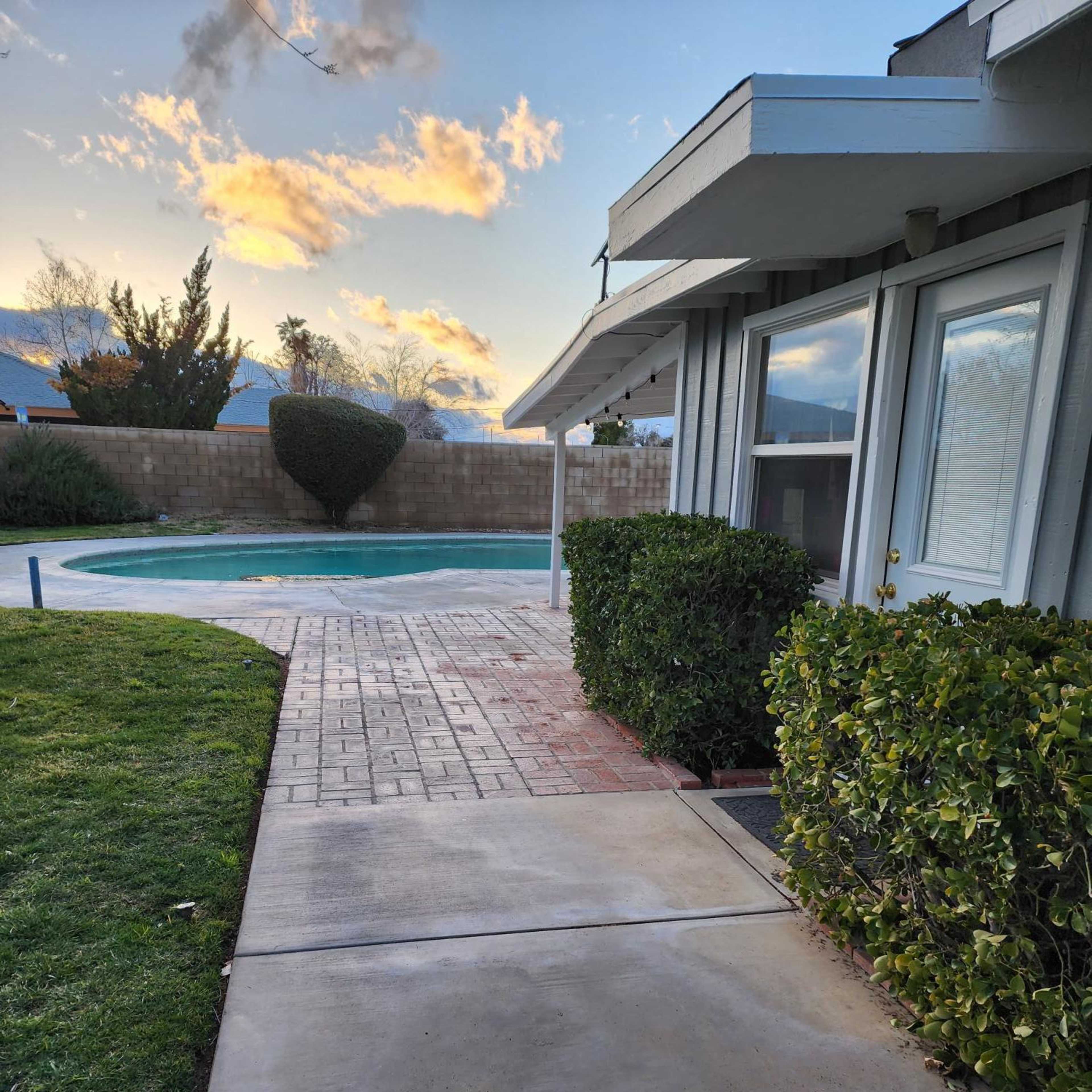 A concrete pathway leads to a pool surrounded by grass and hedges, with a partially cloudy sky visible in the background.