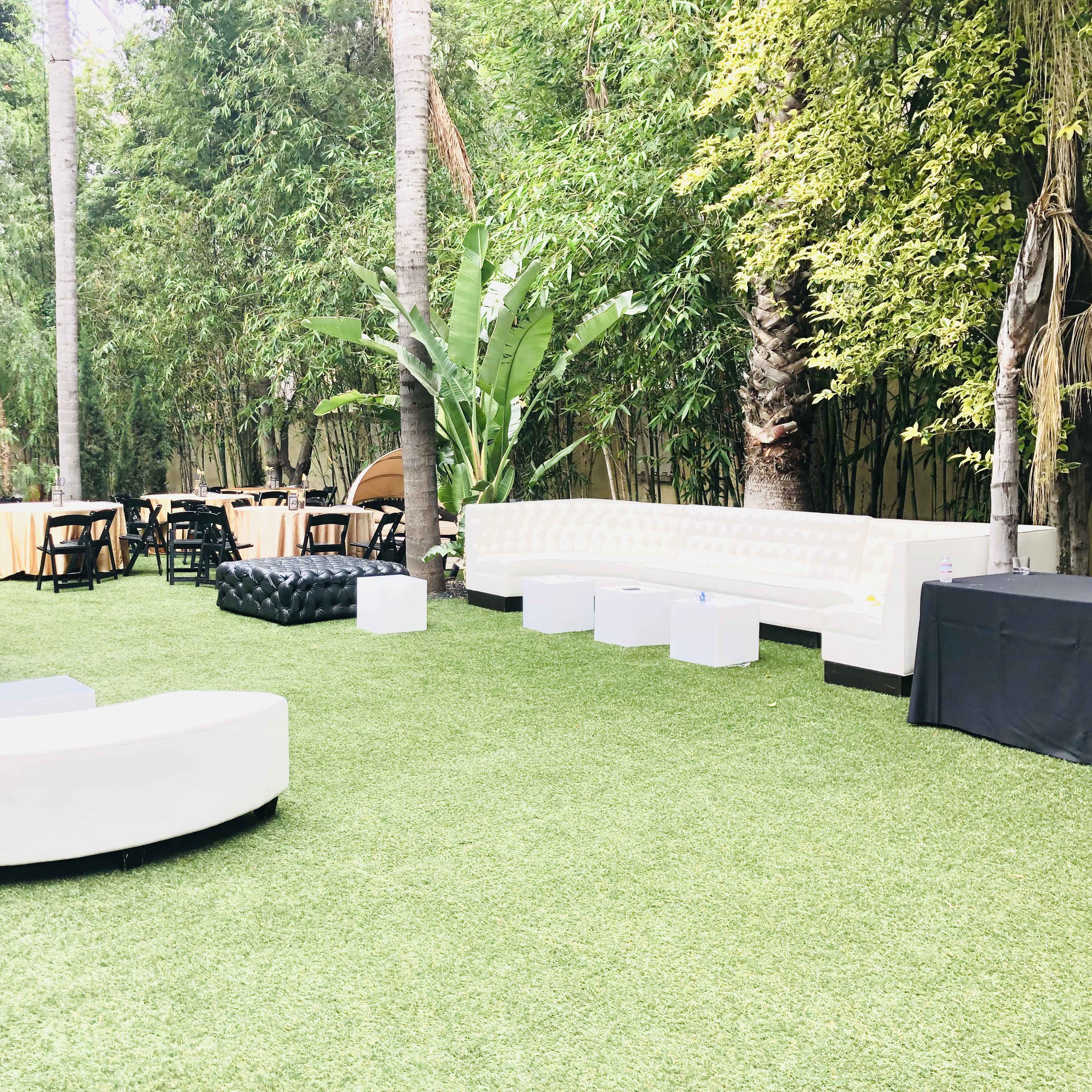 An outdoor event space with white seating, a black ottoman, and round tables set on artificial grass surrounded by greenery.