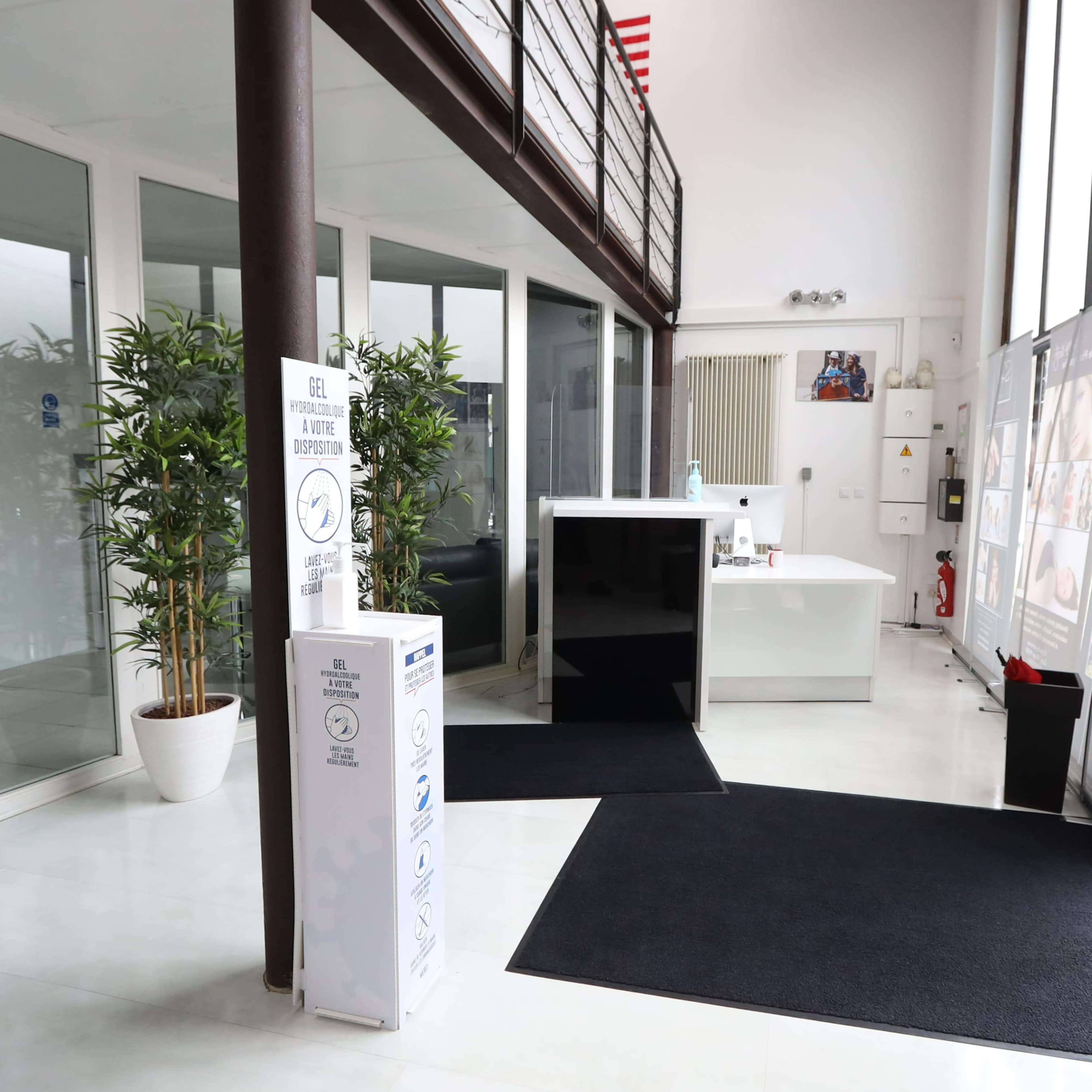 A modern reception area with a white desk, large windows, and plants, featuring a black mat on the floor and informational displays.