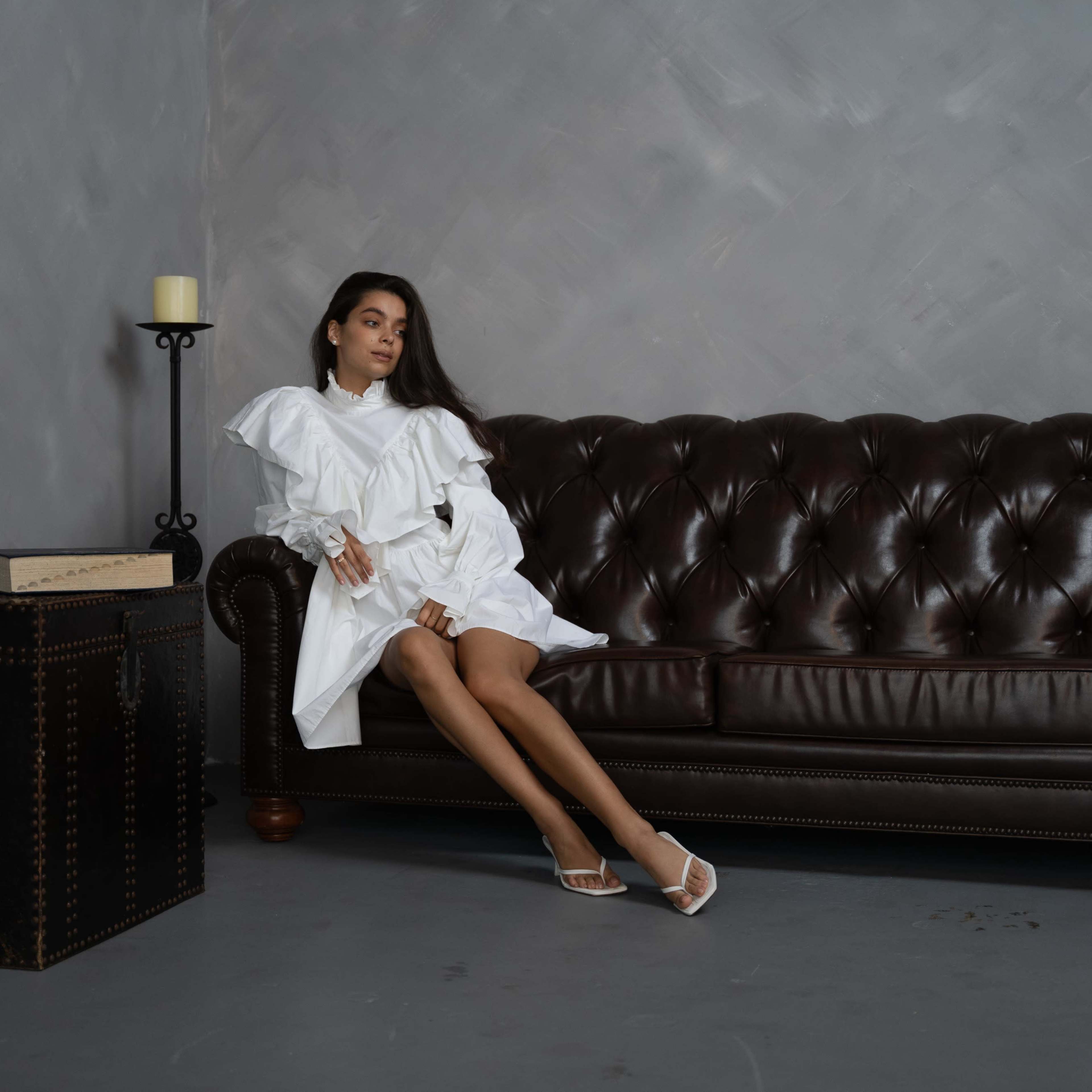 A woman in a white dress sits on a dark brown leather sofa next to a candleholder and a decorative box.