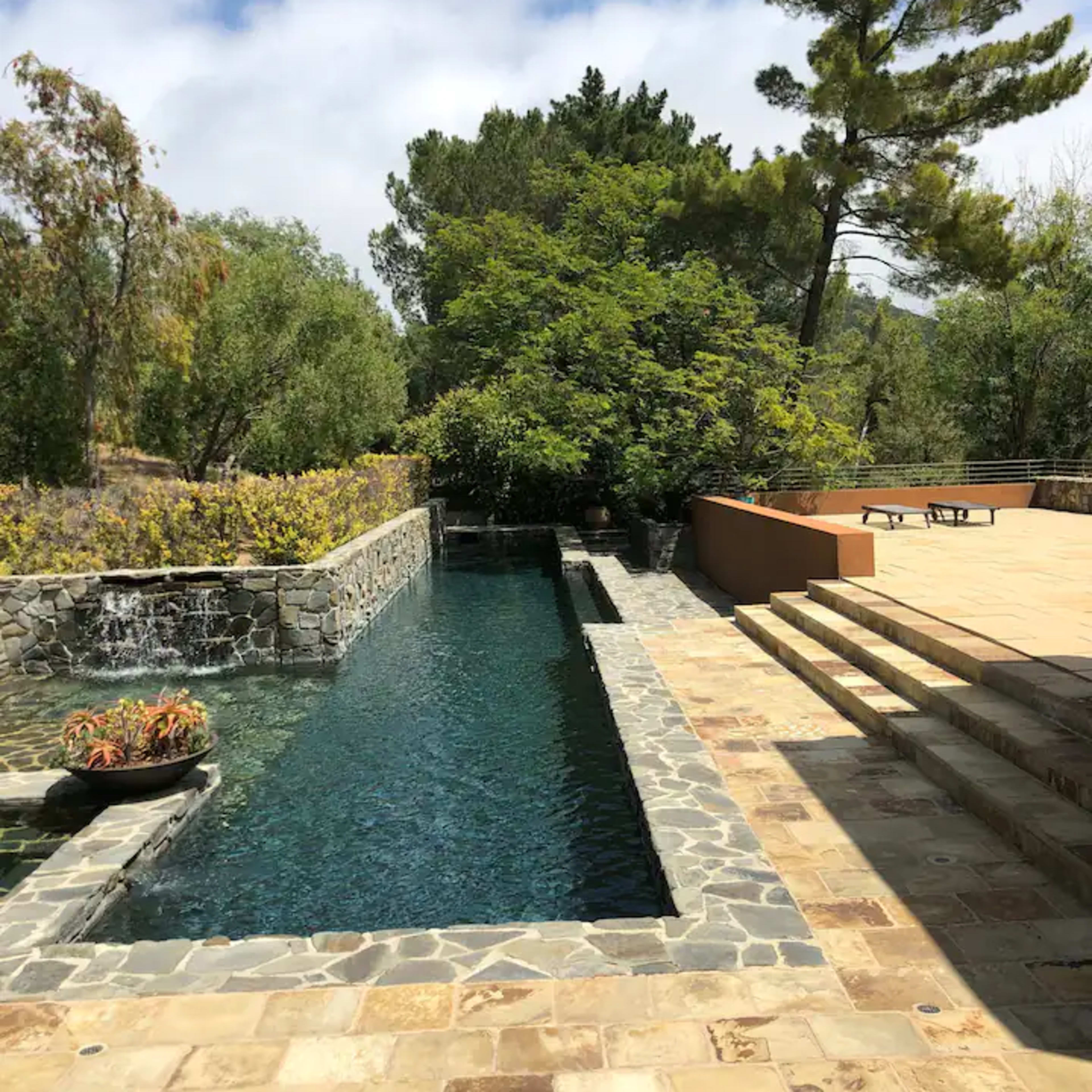 A stone patio with steps leading down to a narrow pool of water bordered by greenery and a small waterfall.