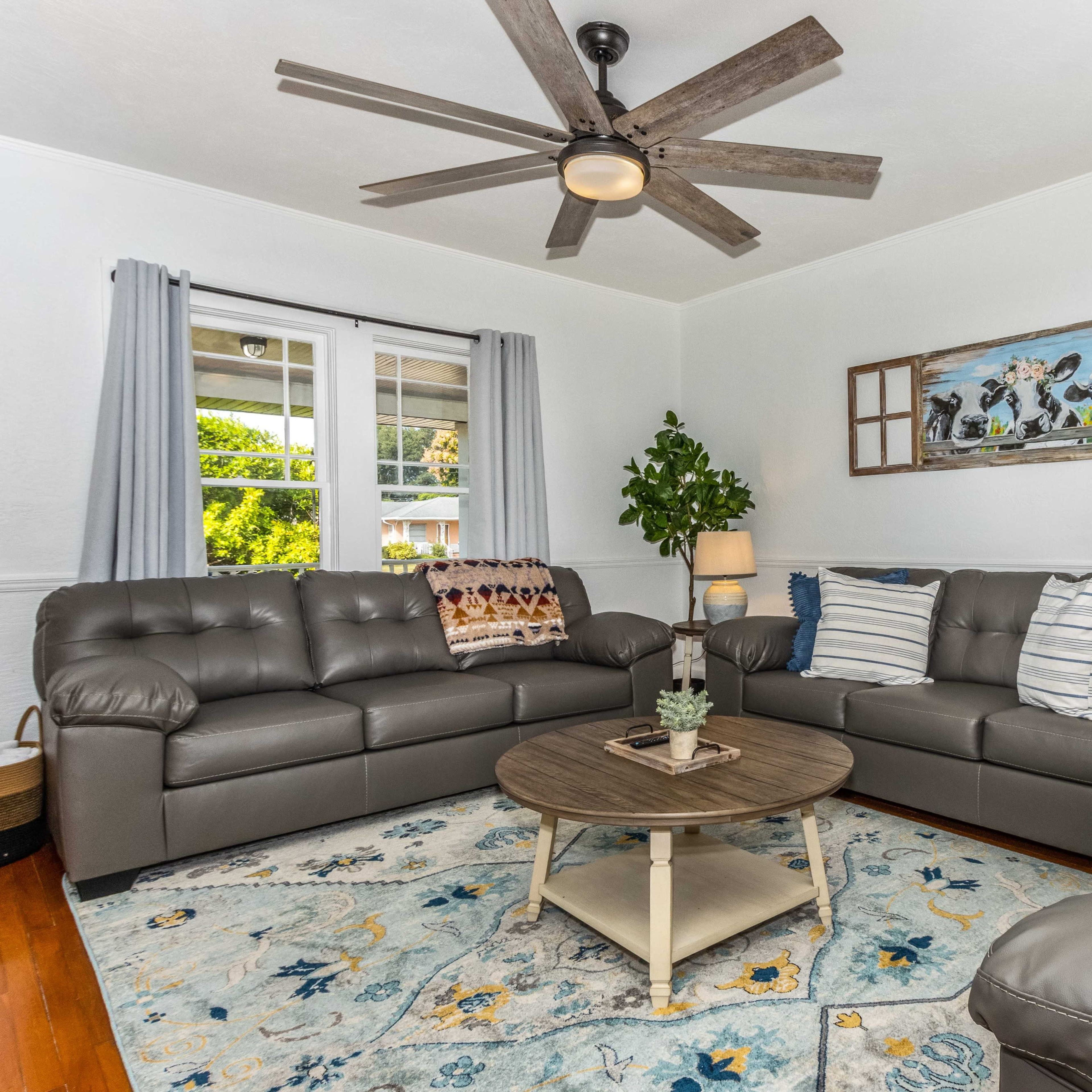A living room features gray leather sofas, a round wooden coffee table, and a decorative blue and yellow area rug under a ceiling fan.
