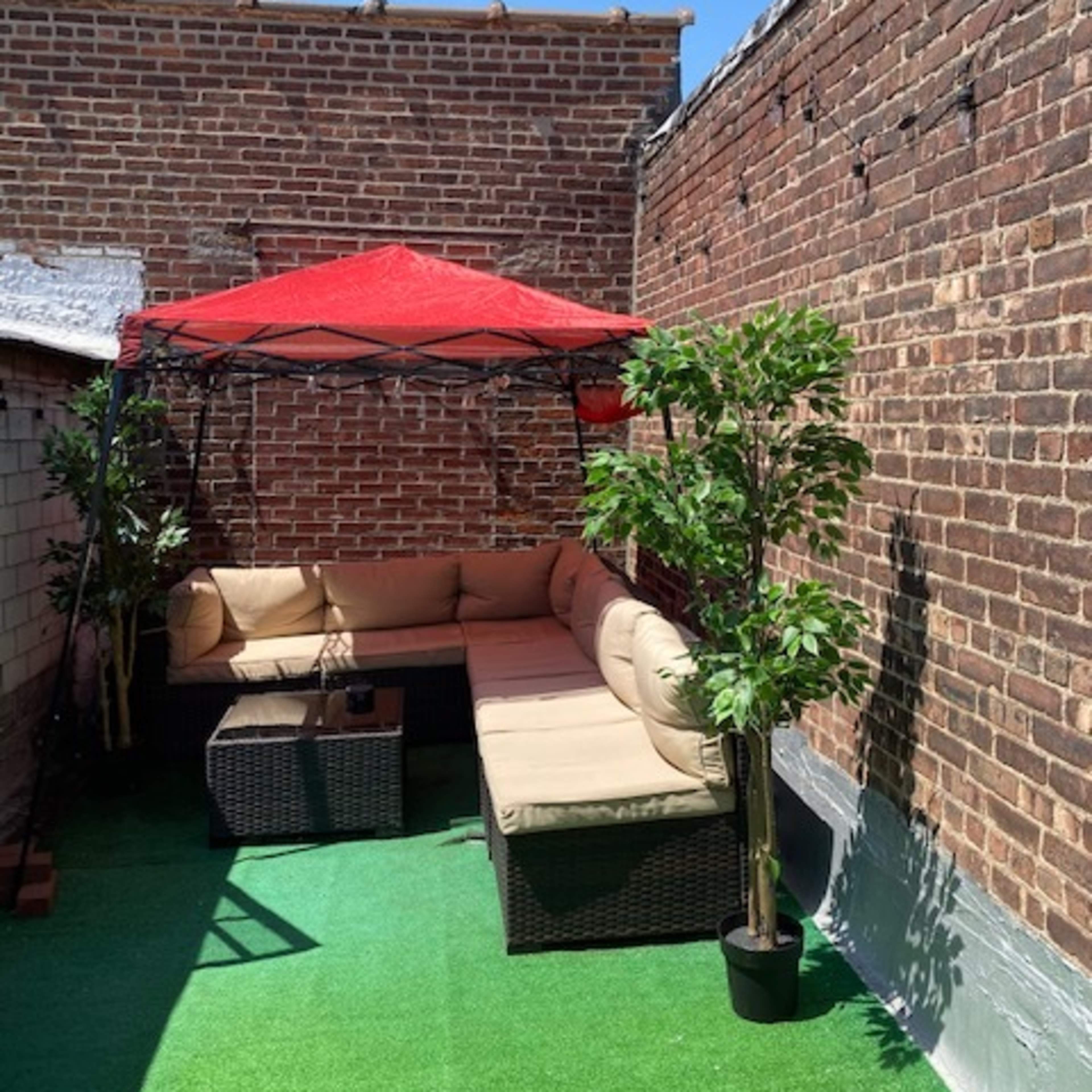 A rooftop terrace features a red canopy over a beige sectional sofa, surrounded by potted plants and artificial grass.