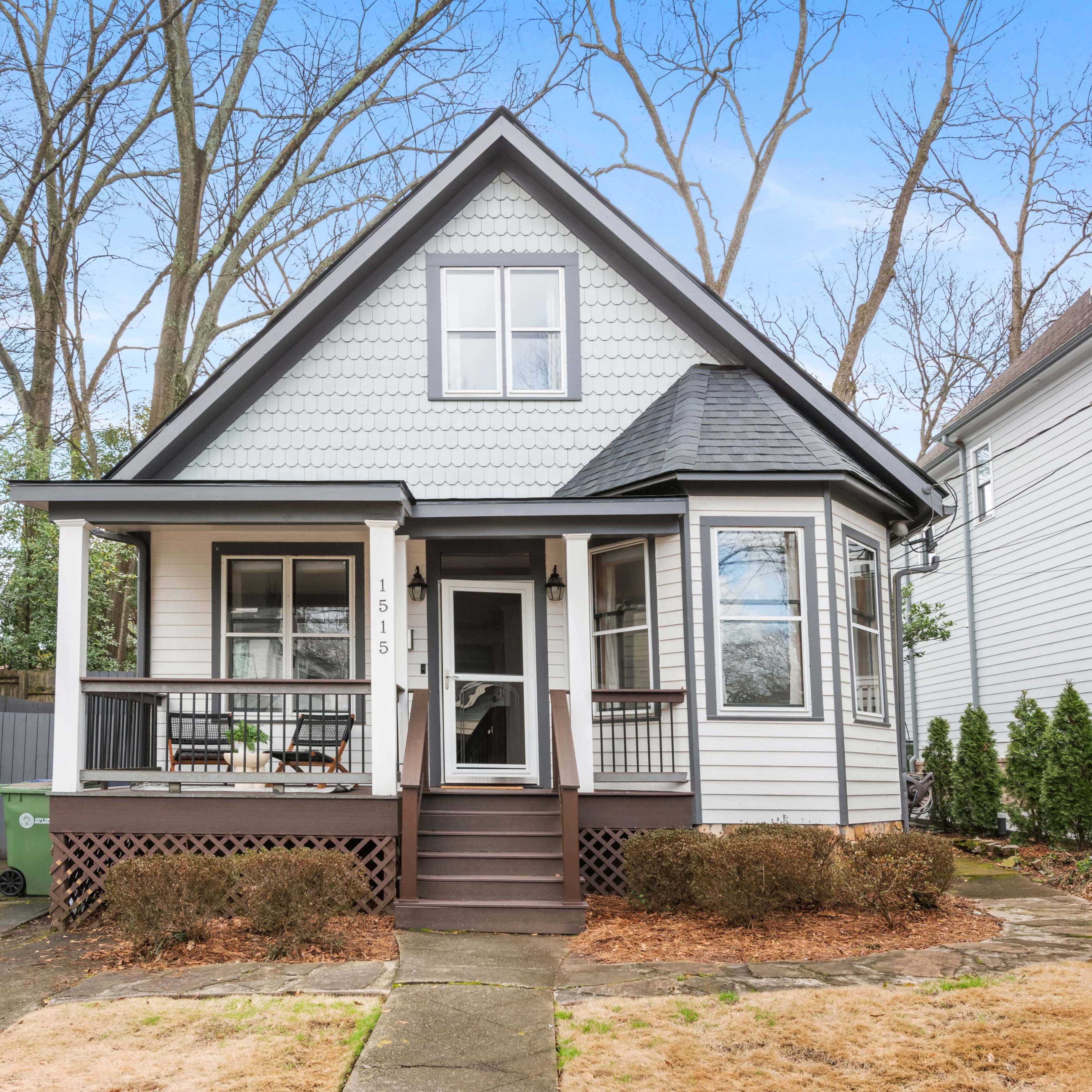 A charming two-story house with a steep roof and a front porch is set among bare trees in a suburban neighborhood.