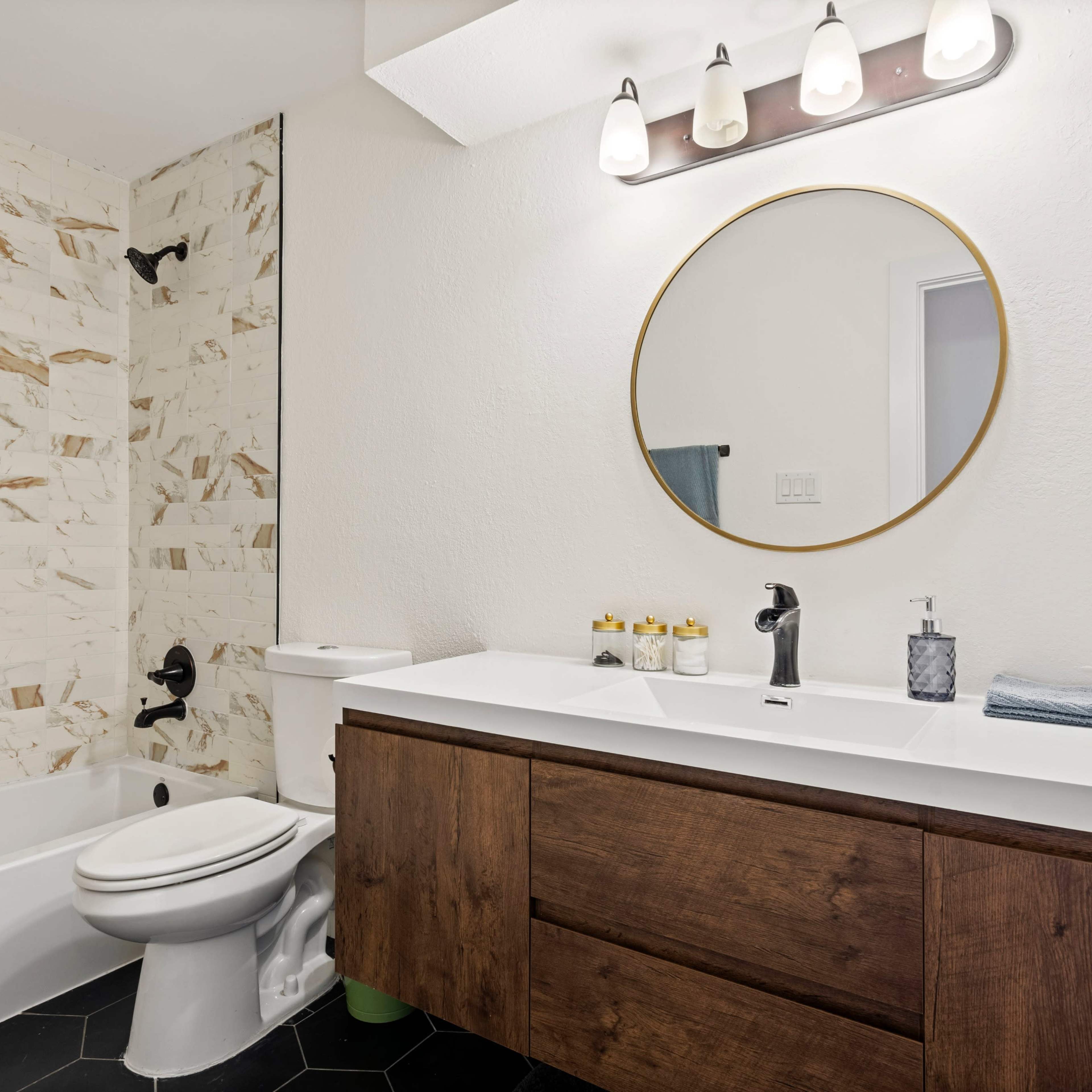 A modern bathroom features a round mirror above a wooden vanity, a bathtub with a tiled wall, and a sleek black faucet.