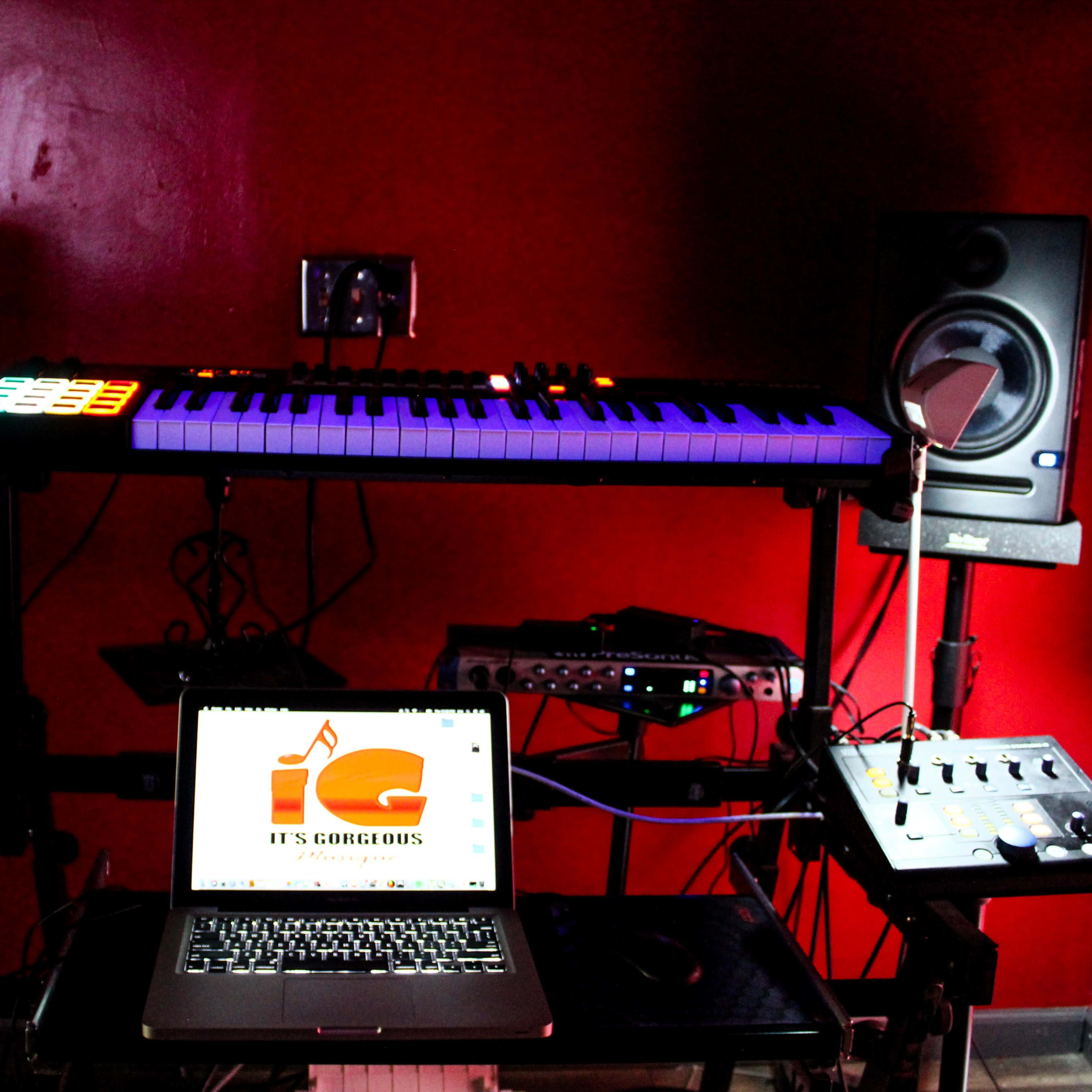 A music production setup featuring a keyboard, a laptop with a logo, and audio equipment against a red wall.