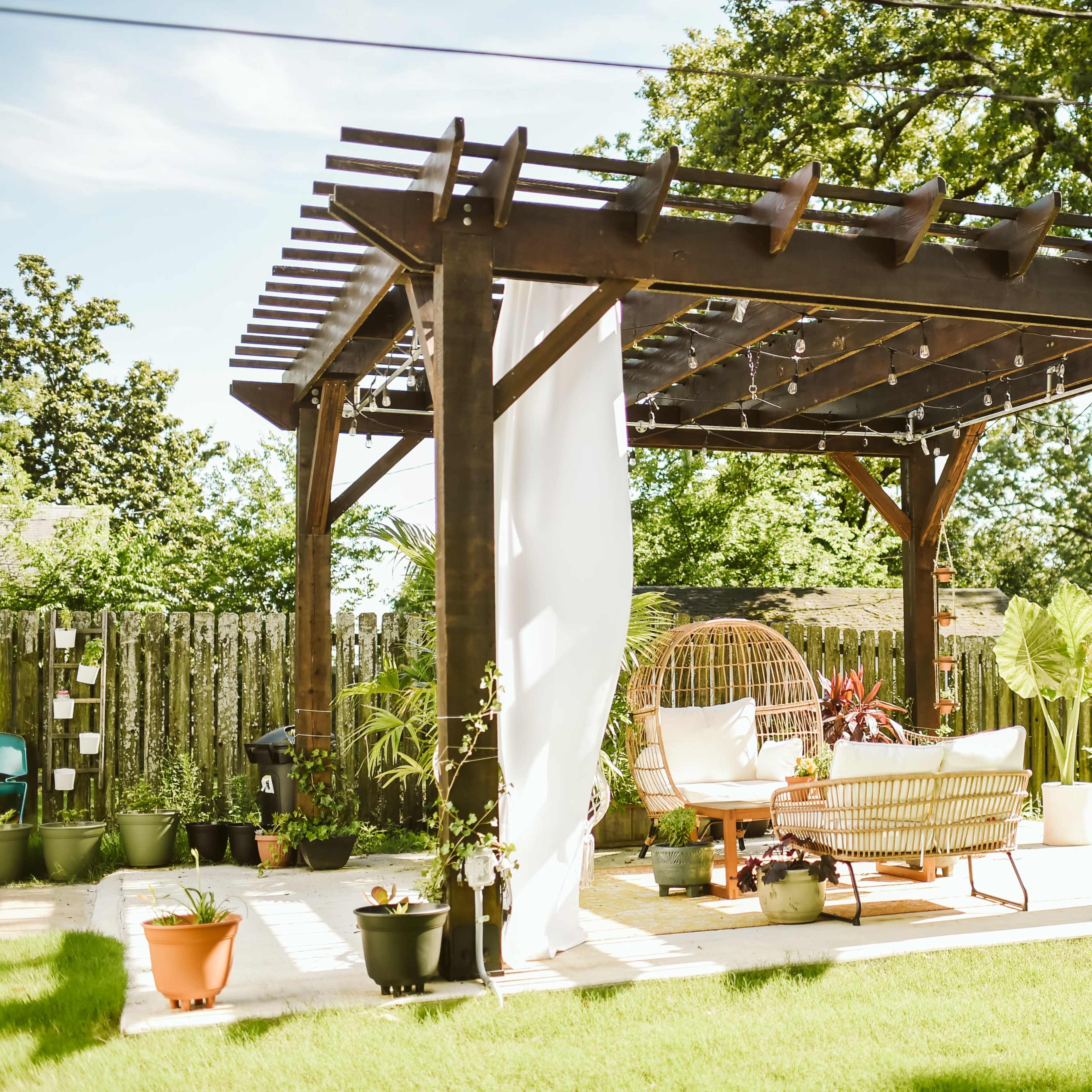 A wooden pergola adorned with hanging lights and sheer curtains shelters a cozy outdoor seating area surrounded by potted plants.