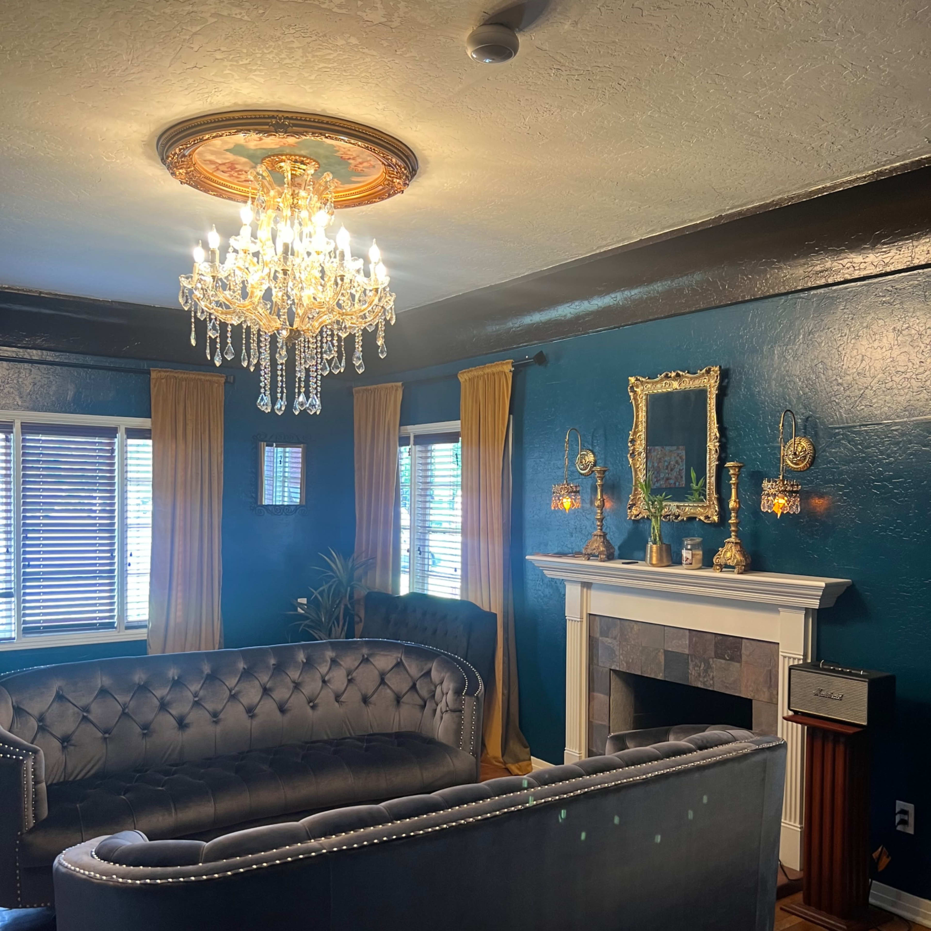 A living room with dark blue walls, a modern chandelier, plush sofas, and a fireplace framed by decorative mirrors and wall sconces.