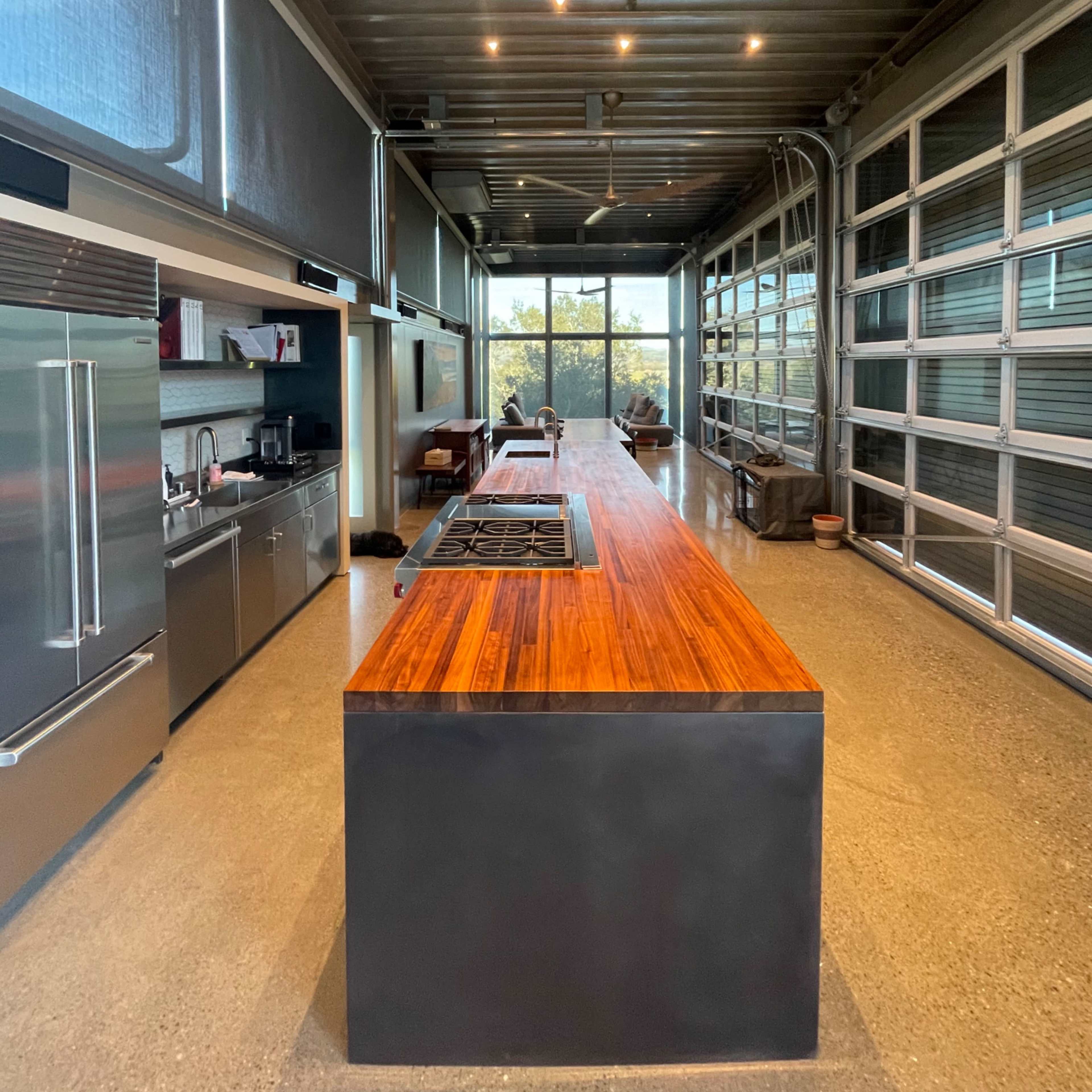 A modern kitchen with a long wooden island in the center, stainless steel appliances along one side, and large windows on the opposite wall.