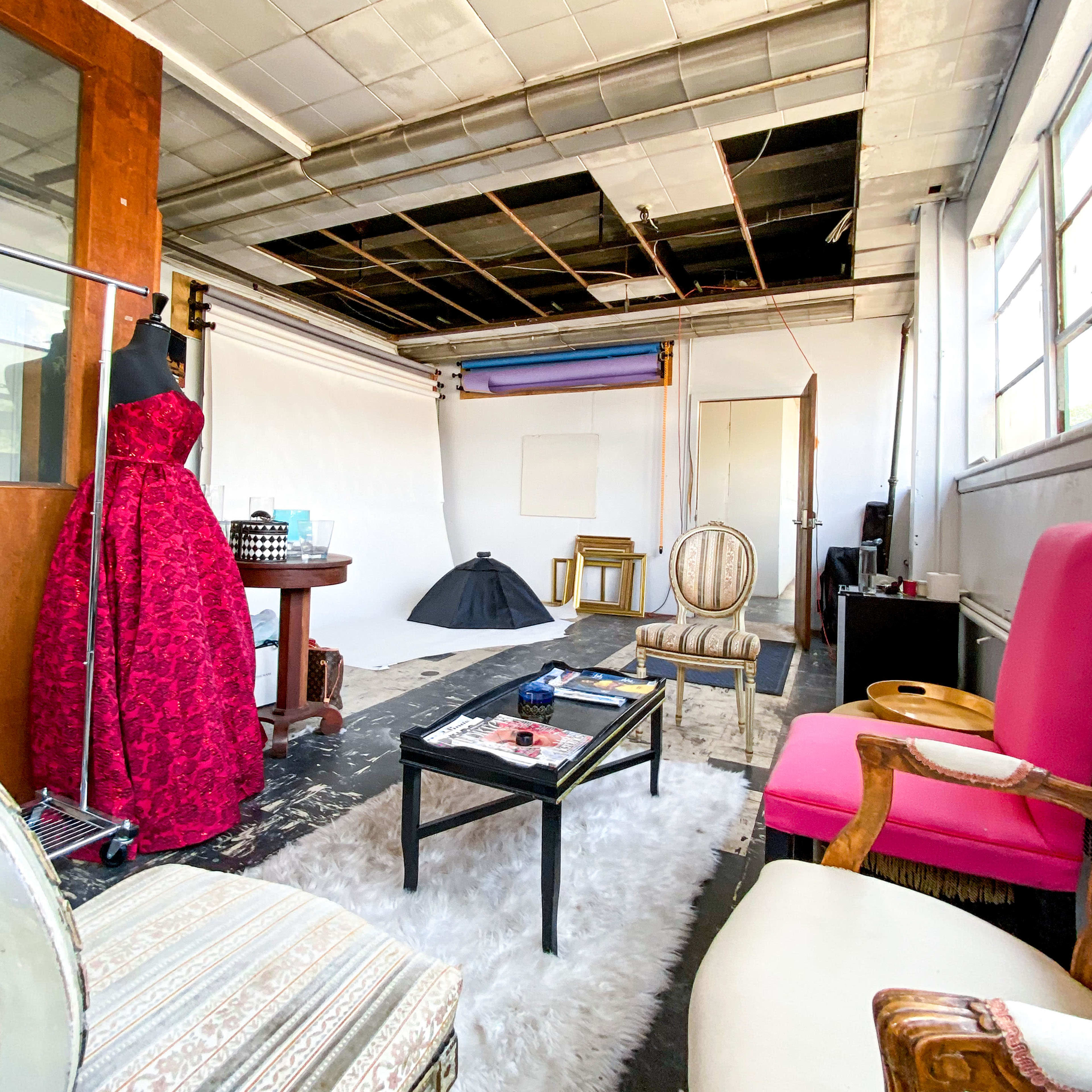 A spacious studio with exposed ceiling tiles, a large pink chair, a vintage table with a mannequin dressed in a red gown, and various props arranged around a white backdrop.