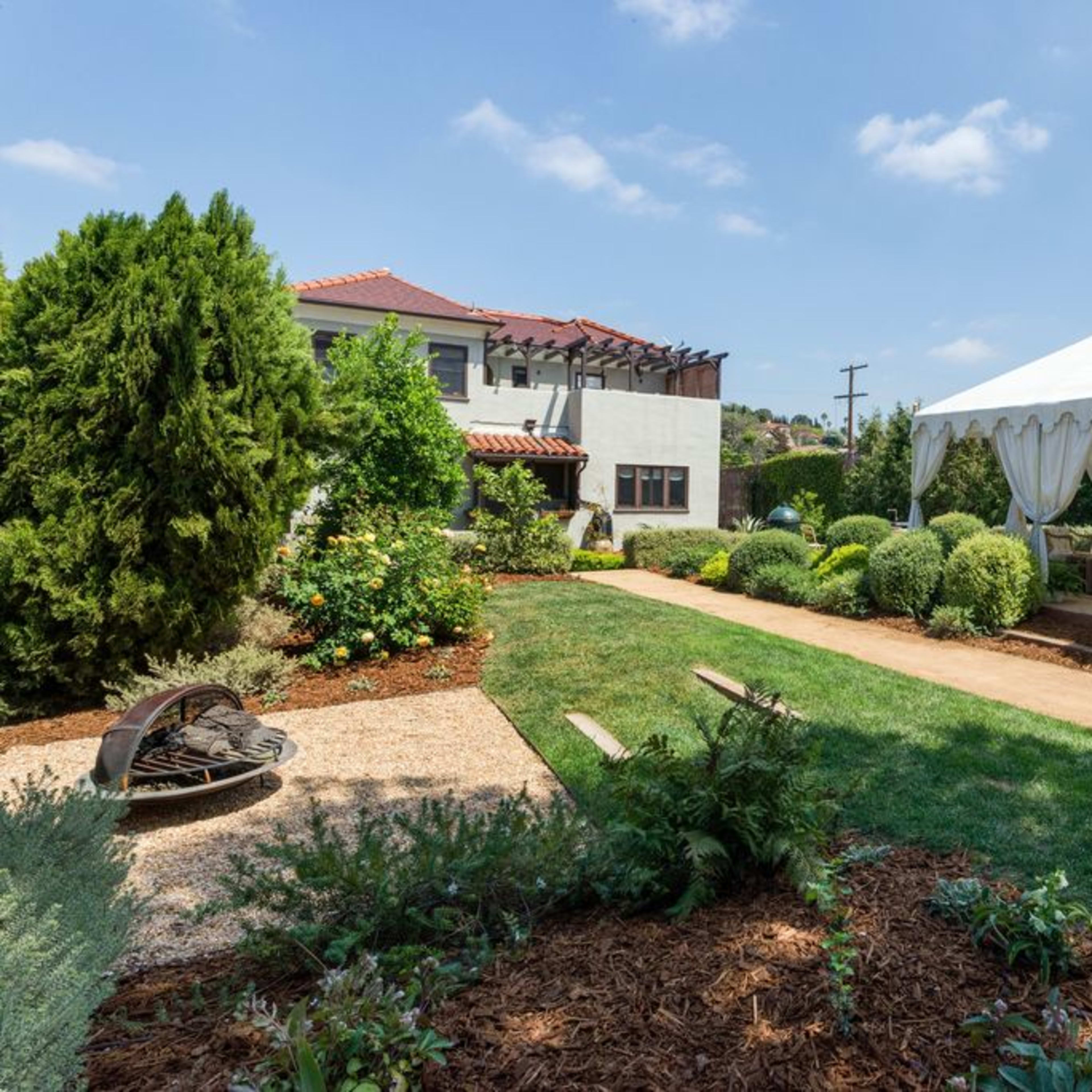 A well-maintained garden features a white tent on a grassy area surrounded by neatly trimmed hedges and plants, with a house visible in the background.