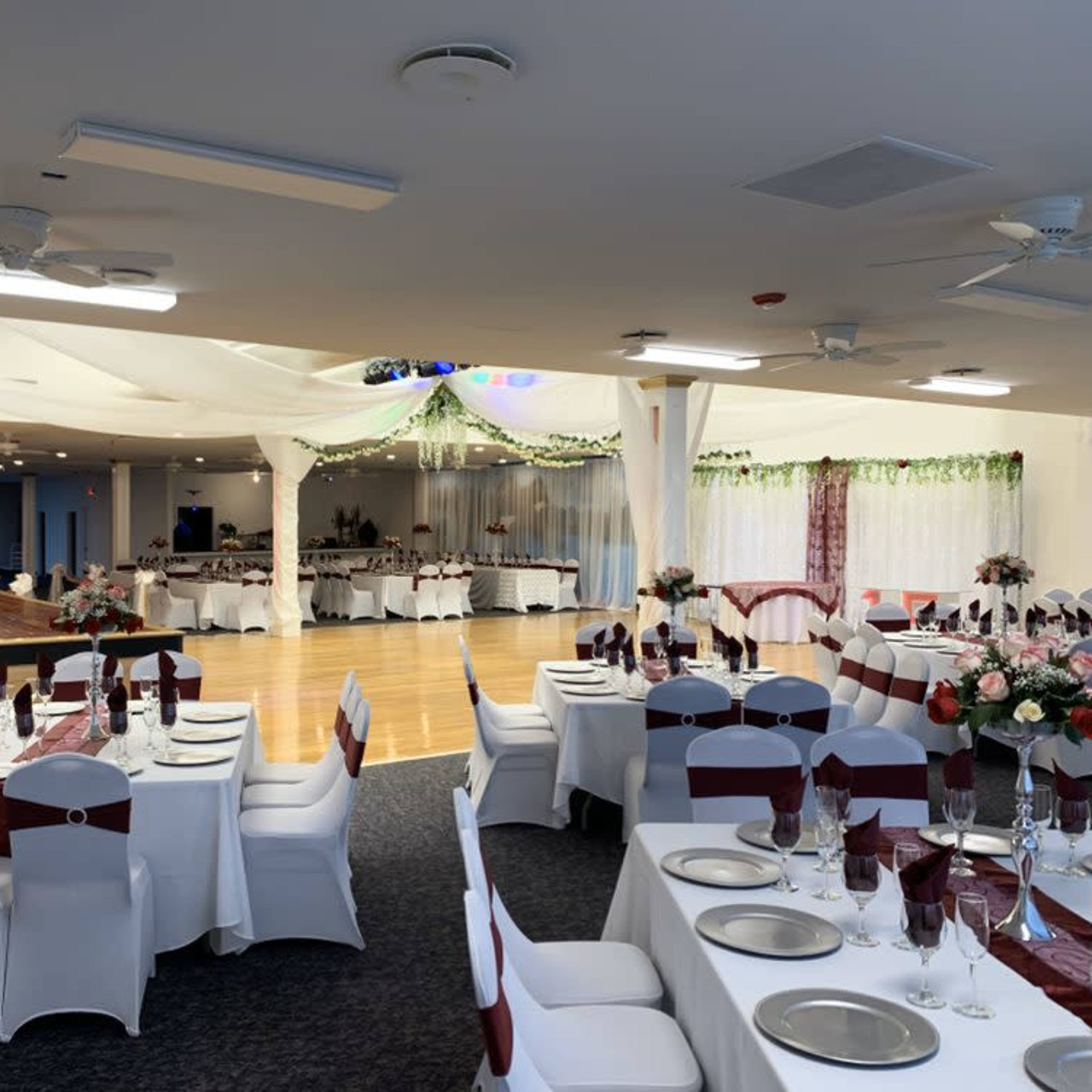 A banquet hall is set up for an event, featuring tables with white tablecloths and red accents, and a dance floor in the background.