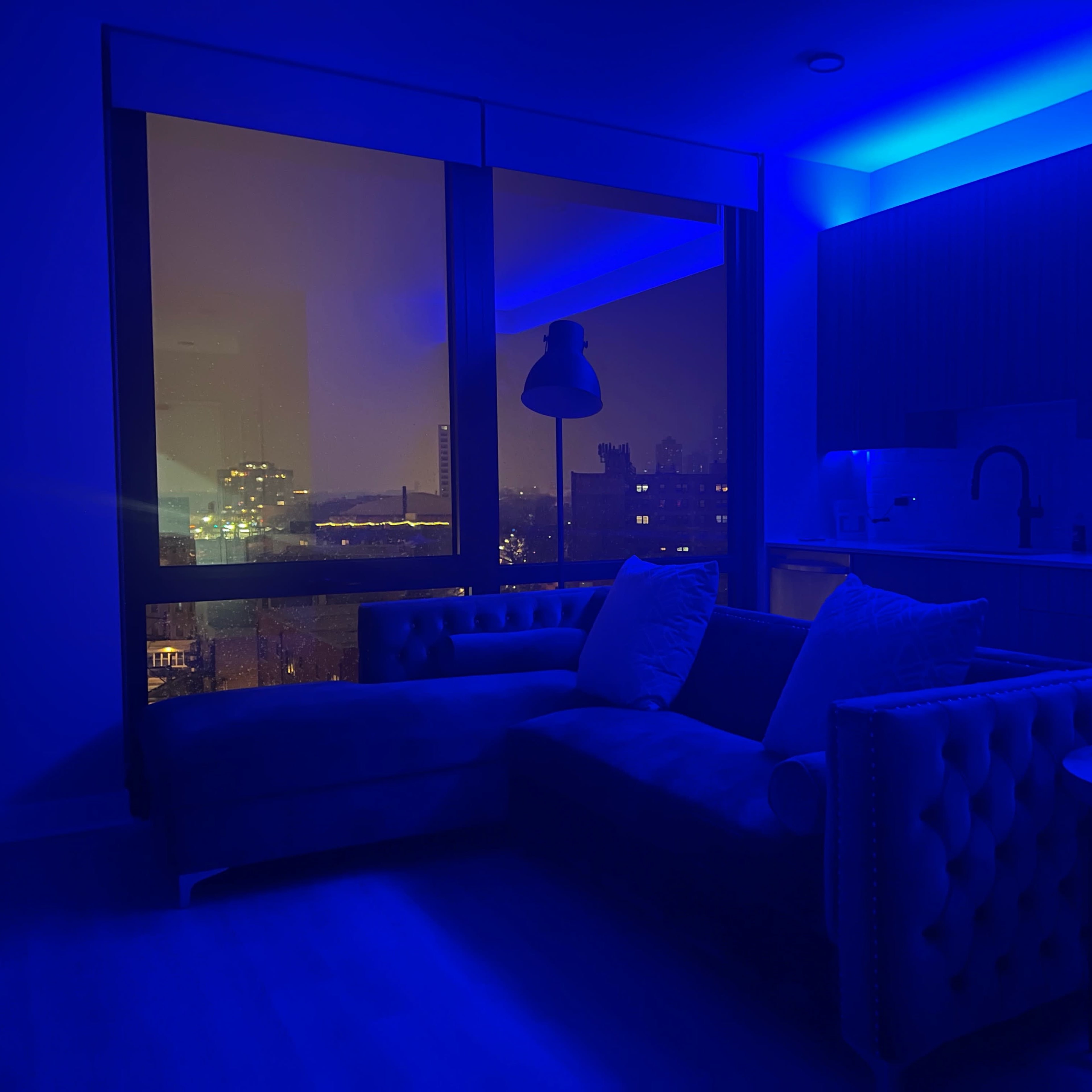 A dimly lit modern apartment features a blue-tinted sofa, a small table with fruit, and a view of a city skyline through large windows.