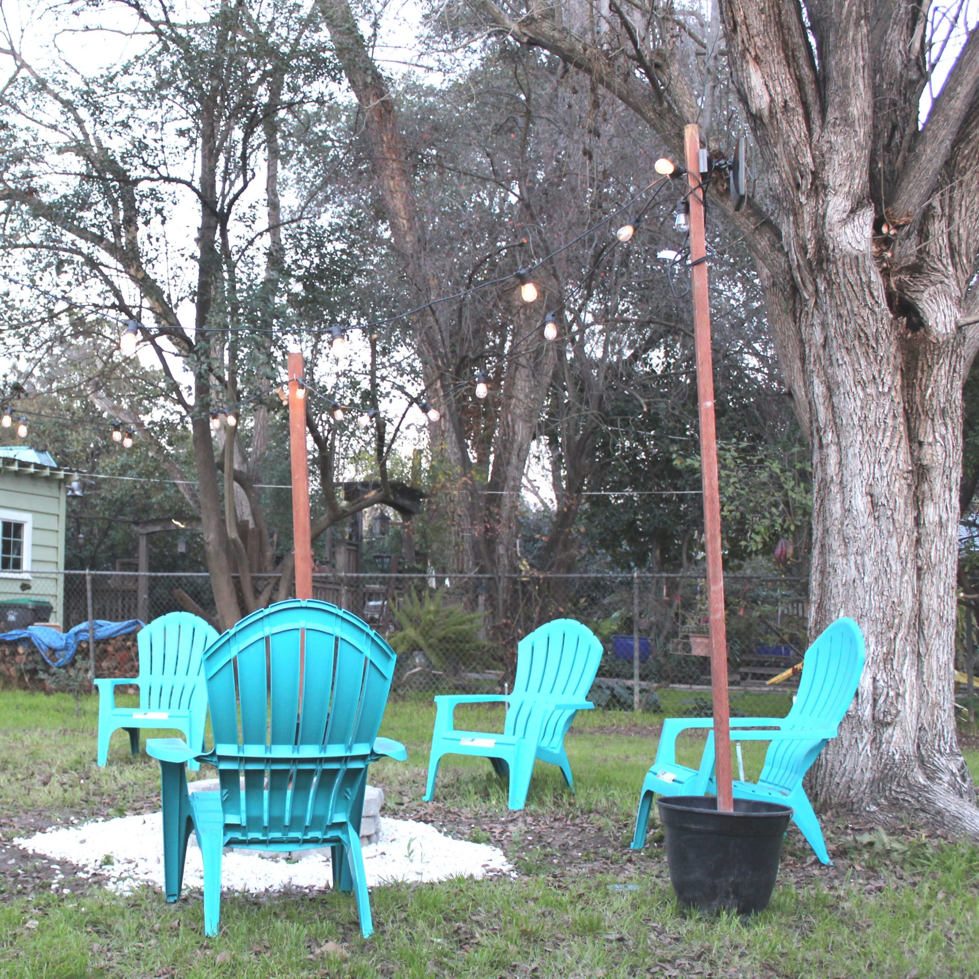 A circular arrangement of turquoise Adirondack chairs surrounds a fire pit in a backyard illuminated by string lights hung between wooden posts.