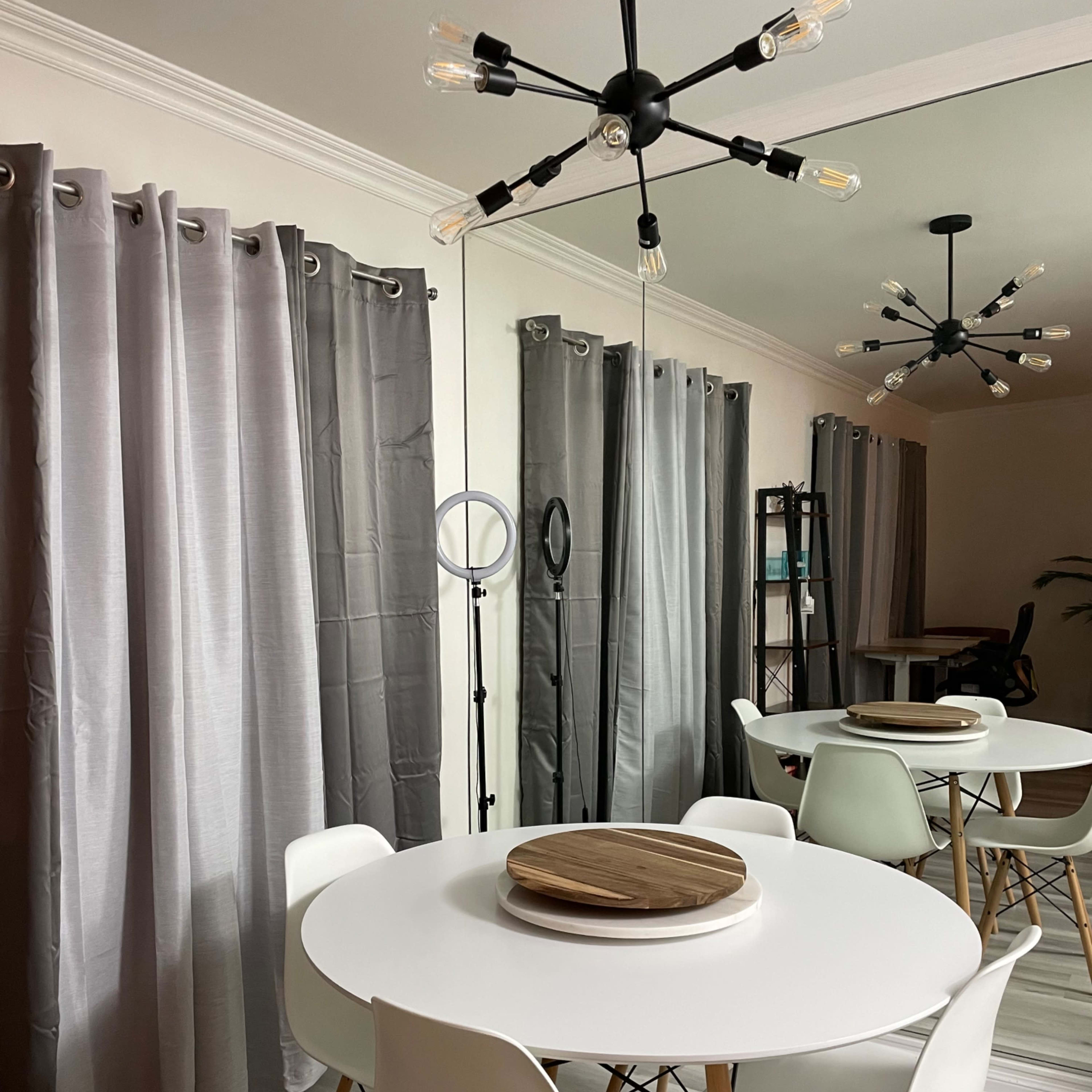 A modern dining area features a round white table surrounded by white chairs, with gray curtains and a geometric light fixture overhead.