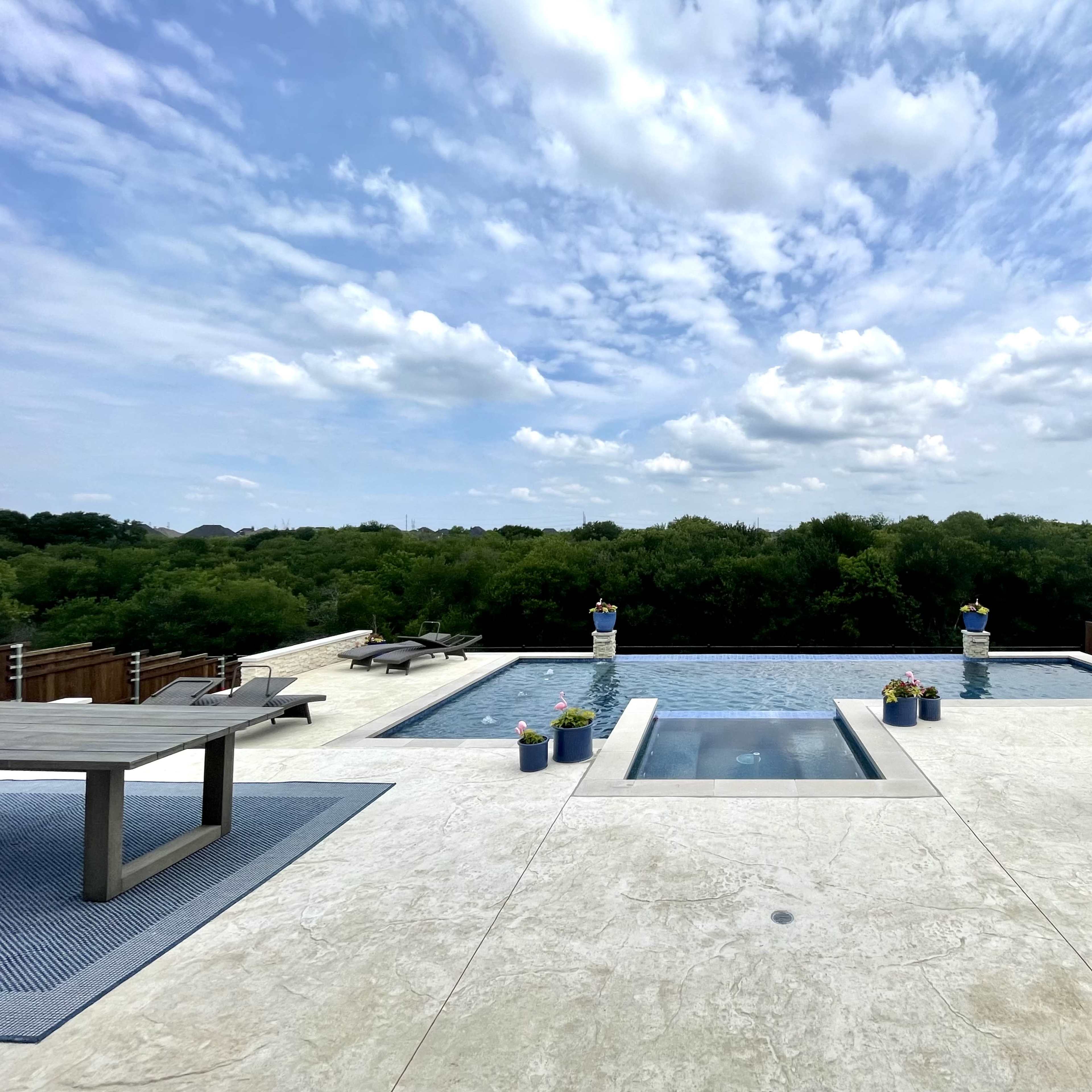 A modern outdoor space features a rectangular pool surrounded by a deck, chairs, and potted plants, overlooking a lush green landscape under a cloudy sky.