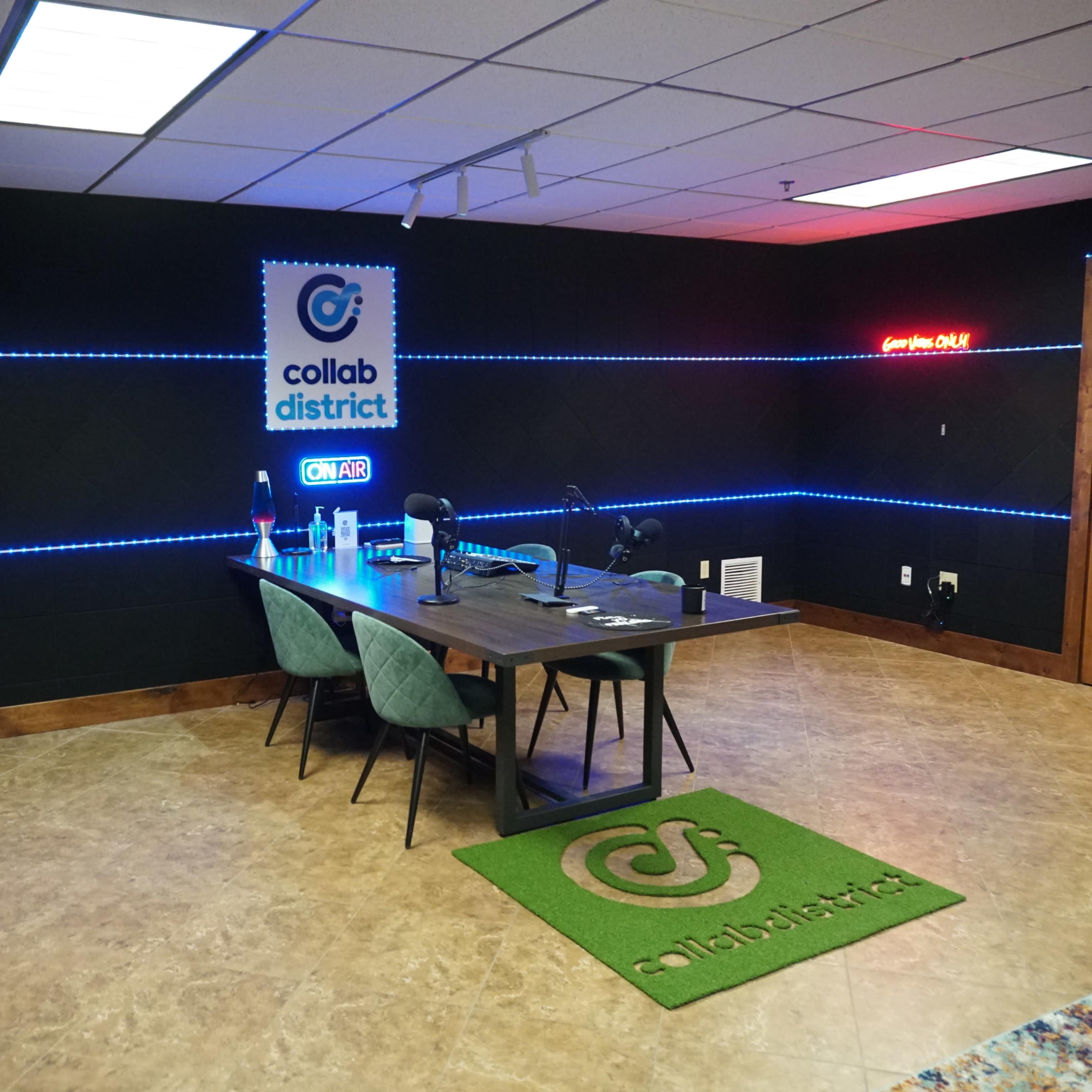 A modern podcasting studio with a wooden table, green chairs, illuminated walls, and a sign that reads "collab district ON AIR."