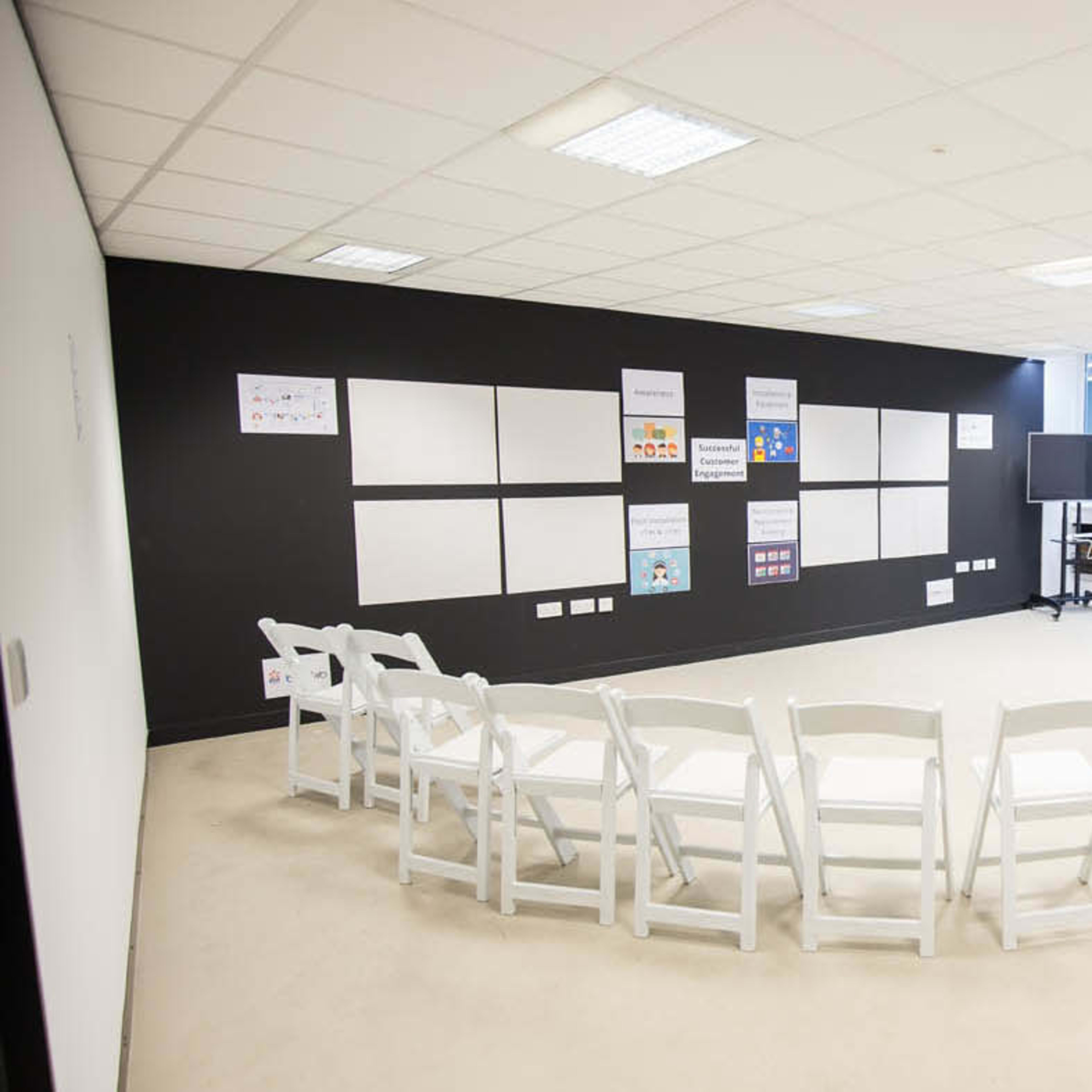 A meeting room features white chairs arranged in a circle facing a wall with various posters and a table in the corner.