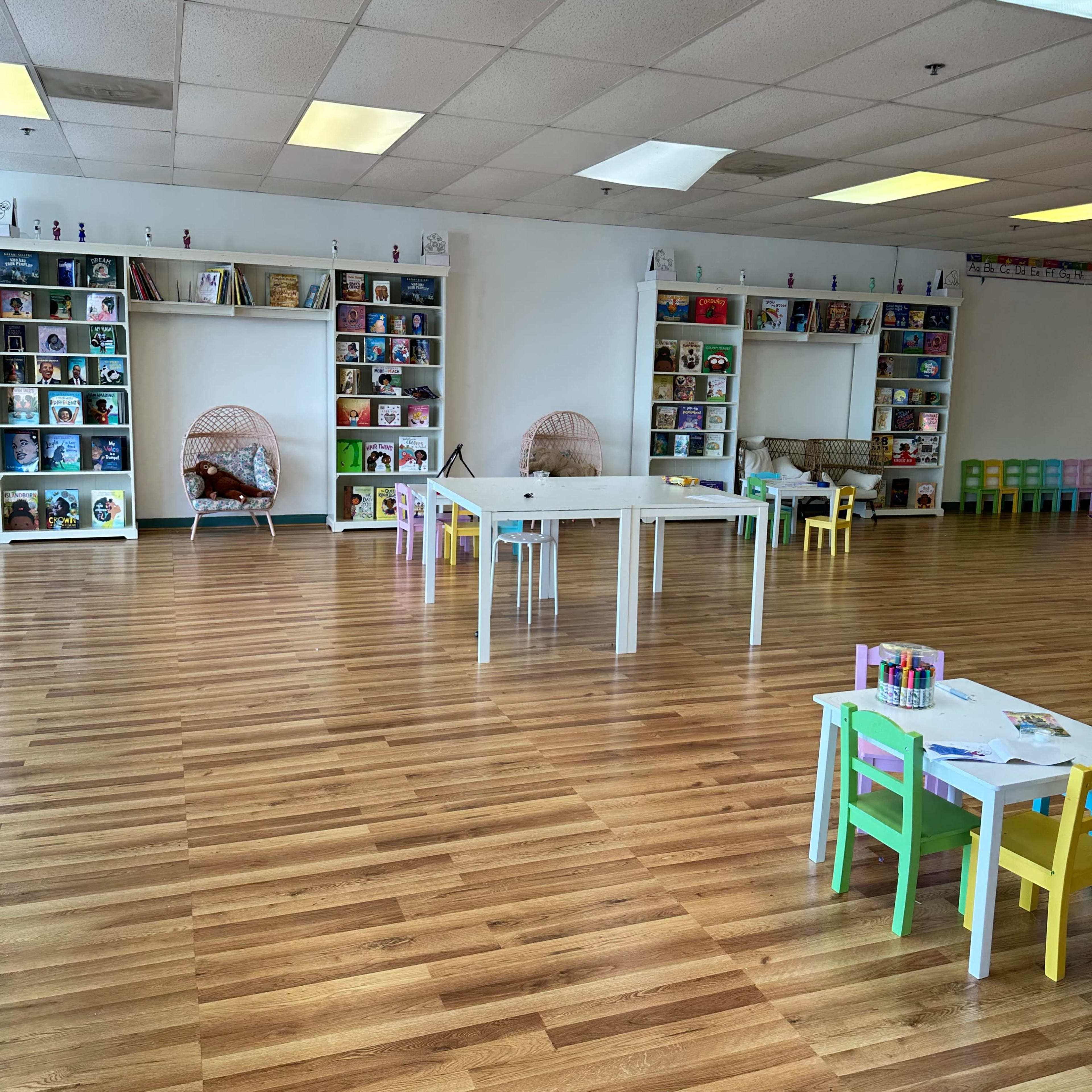 A spacious children's reading area with white shelves filled with books and several small tables and chairs arranged on a wooden floor.