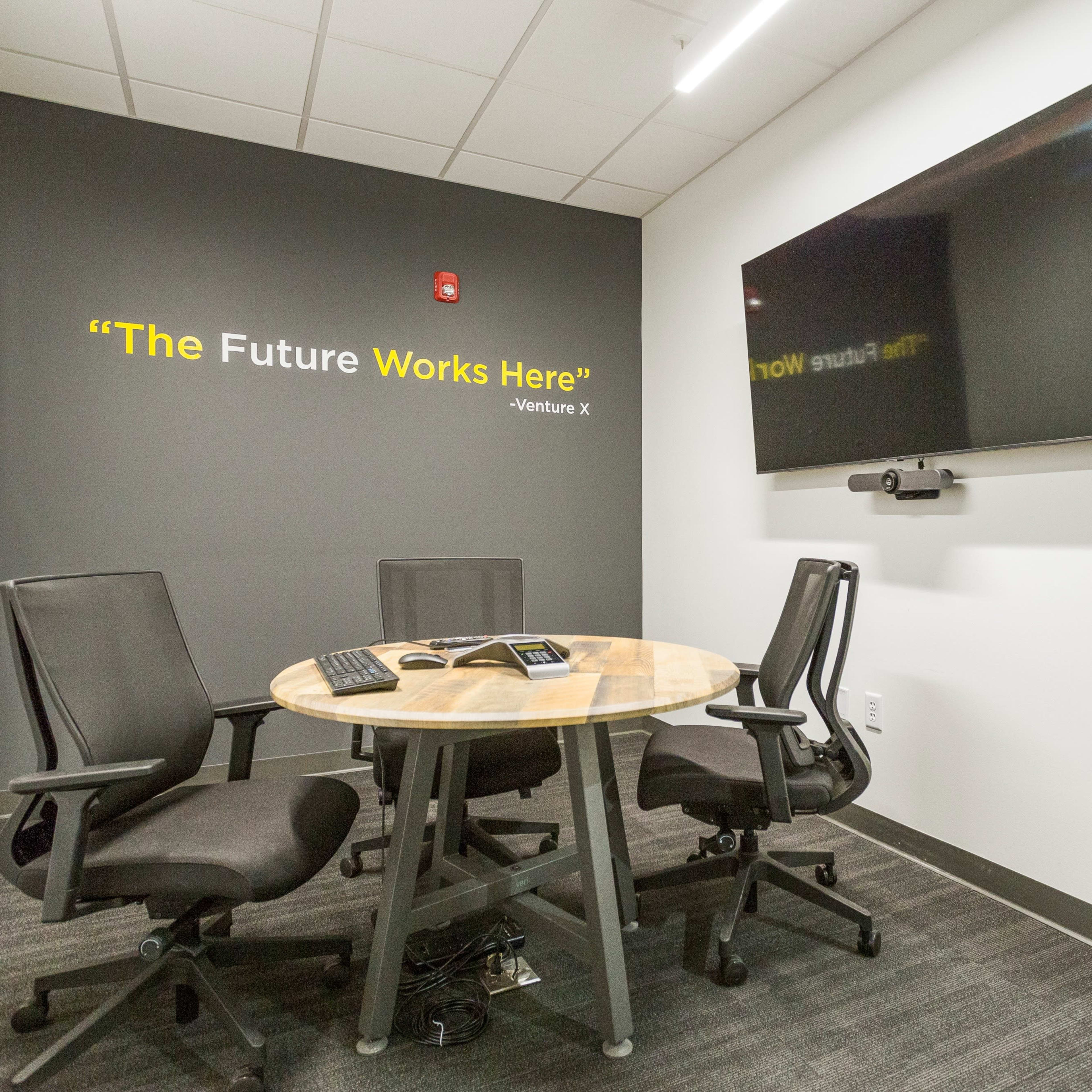 A small conference room features a round wooden table with three black chairs, a wall-mounted television, and a quote displayed on the wall.