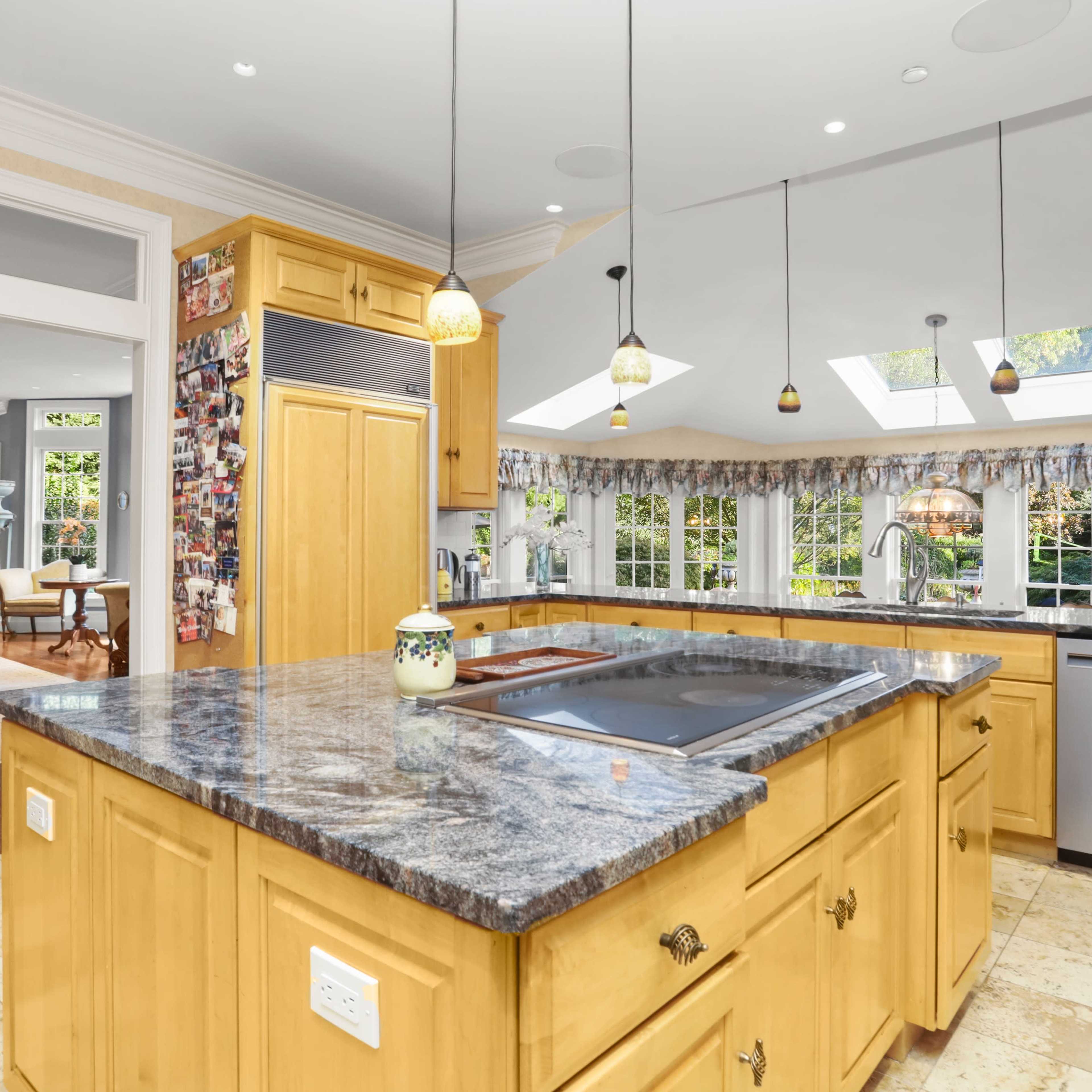 A spacious kitchen with a large island featuring a dark countertop, wooden cabinetry, and pendant lighting above, connected to a living area visible in the background.