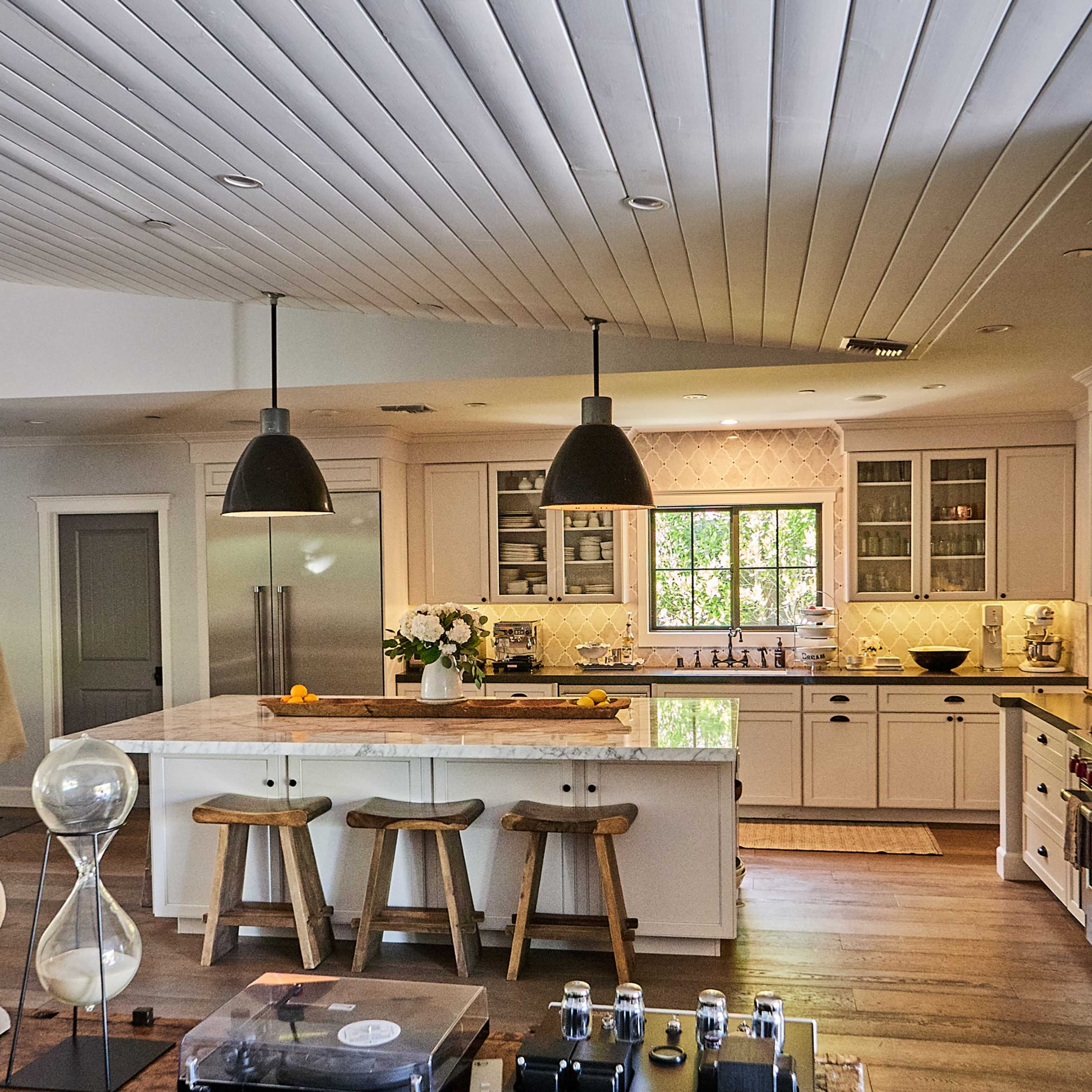 A modern kitchen with white cabinets, a central island, three wooden stools, and large windows letting in natural light.