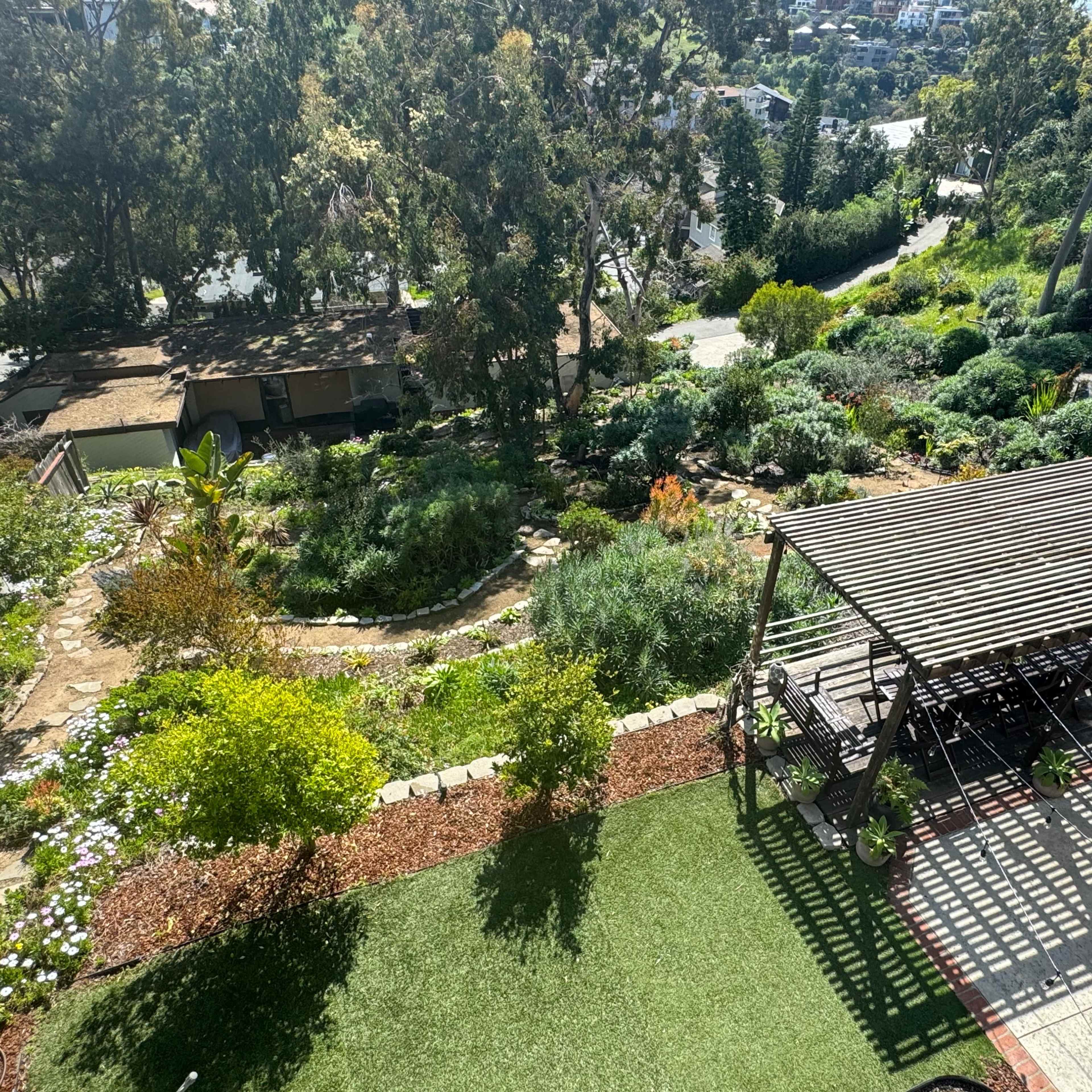 A landscaped garden with a wooden gazebo, stone pathways, and greenery, set against a backdrop of a hillside.
