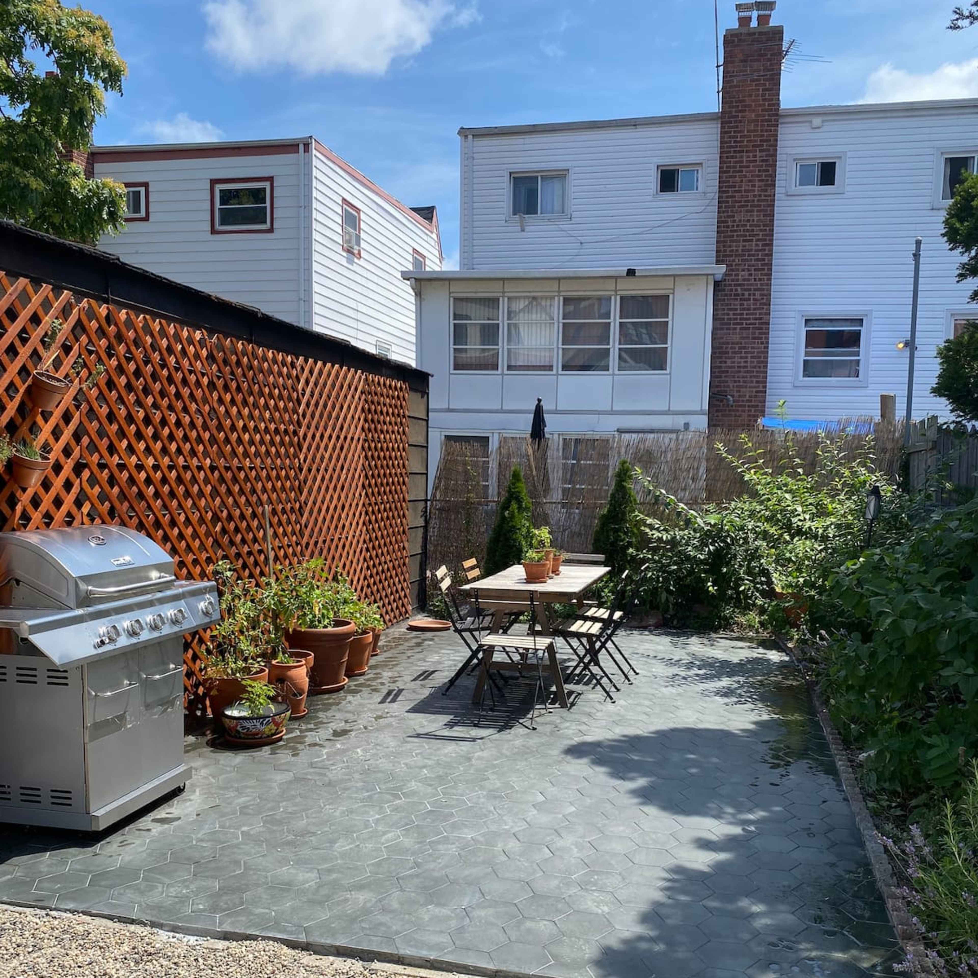 A small outdoor patio area with a table and chairs, a barbecue grill, potted plants, and surrounding wooden fencing.