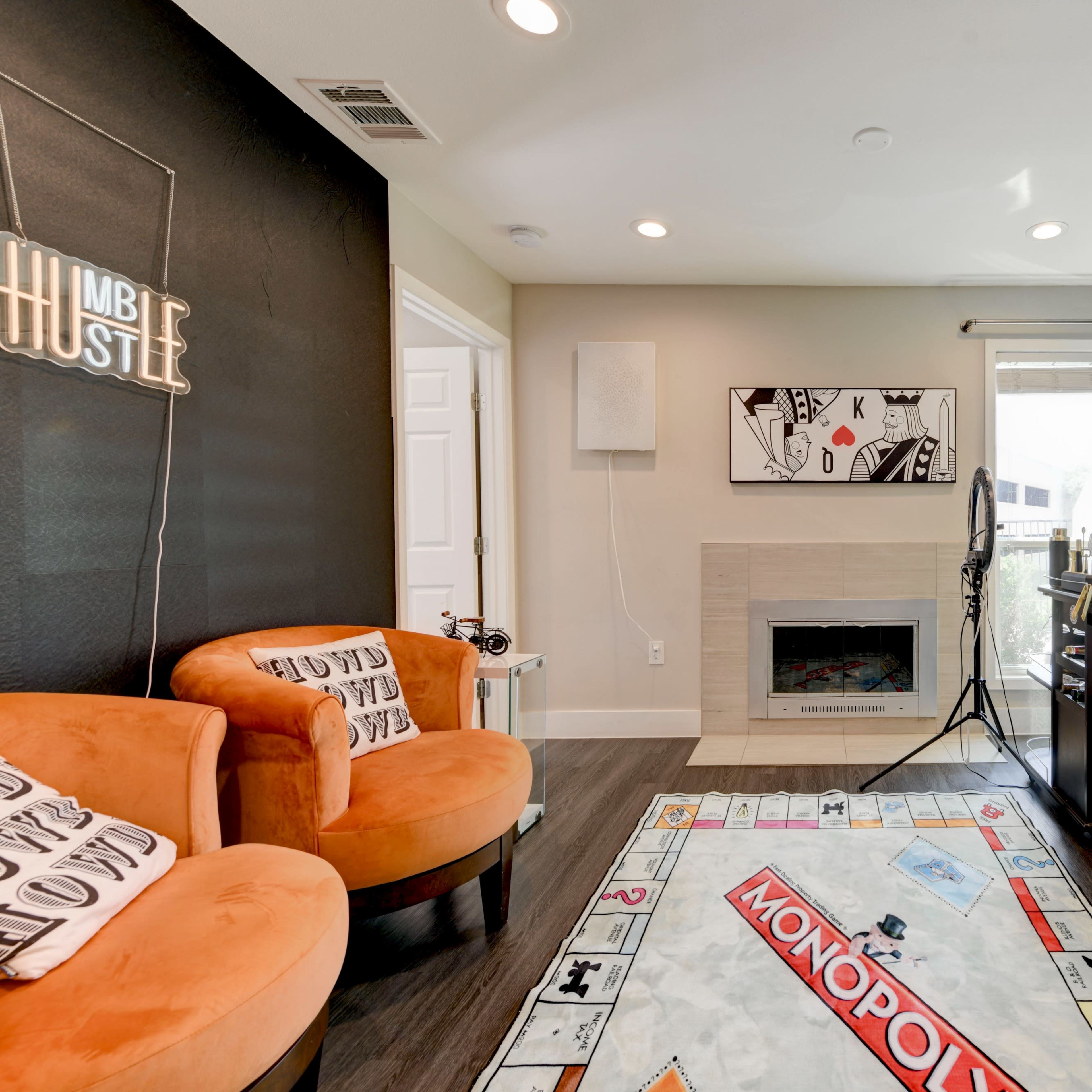 A cozy room featuring a green leafy accent wall, a fireplace, comfortable orange chairs, and a Monopoly-themed rug.