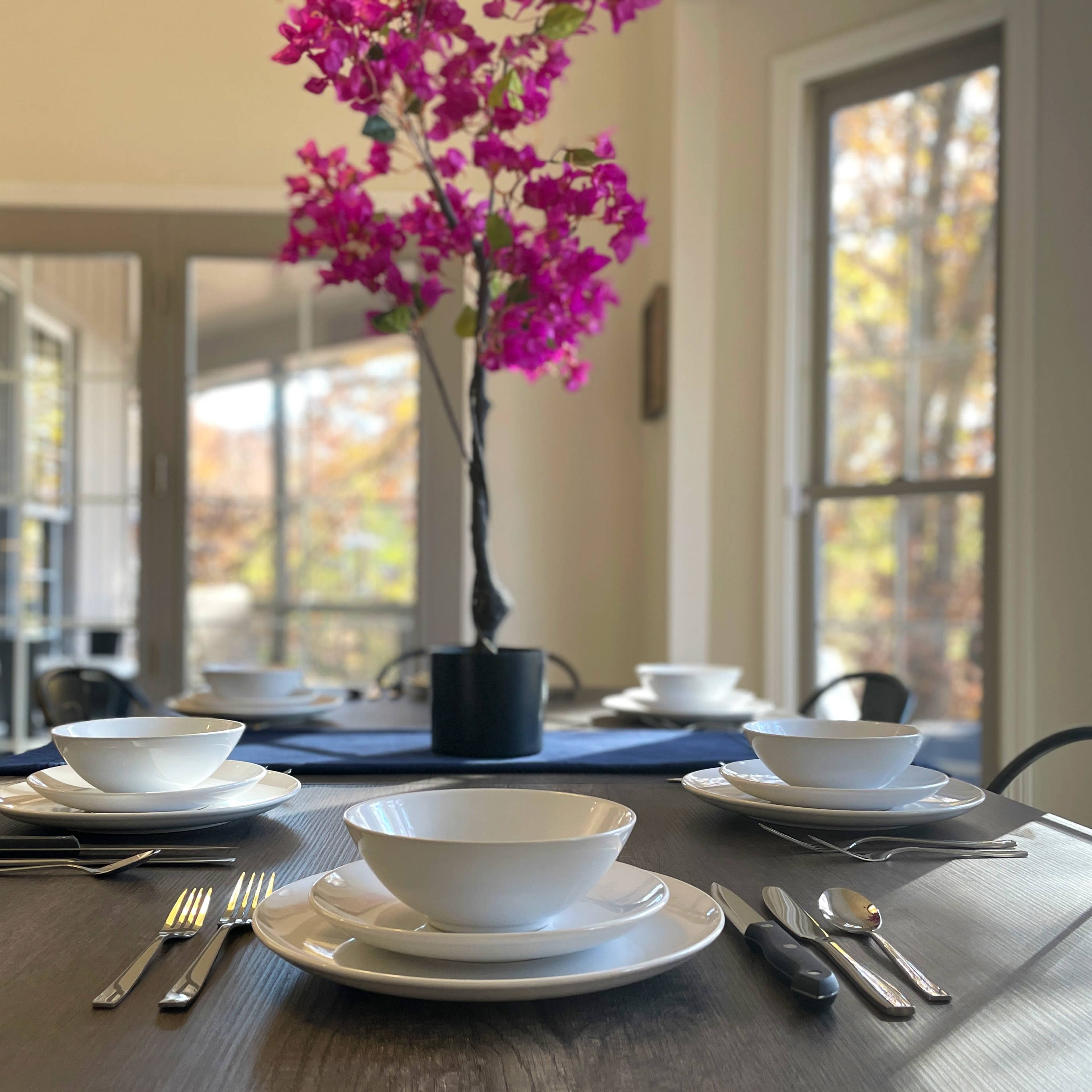 A dining table is set with white bowls and flatware, and a vase of pink flowers is placed in the center, against a backdrop of large windows.