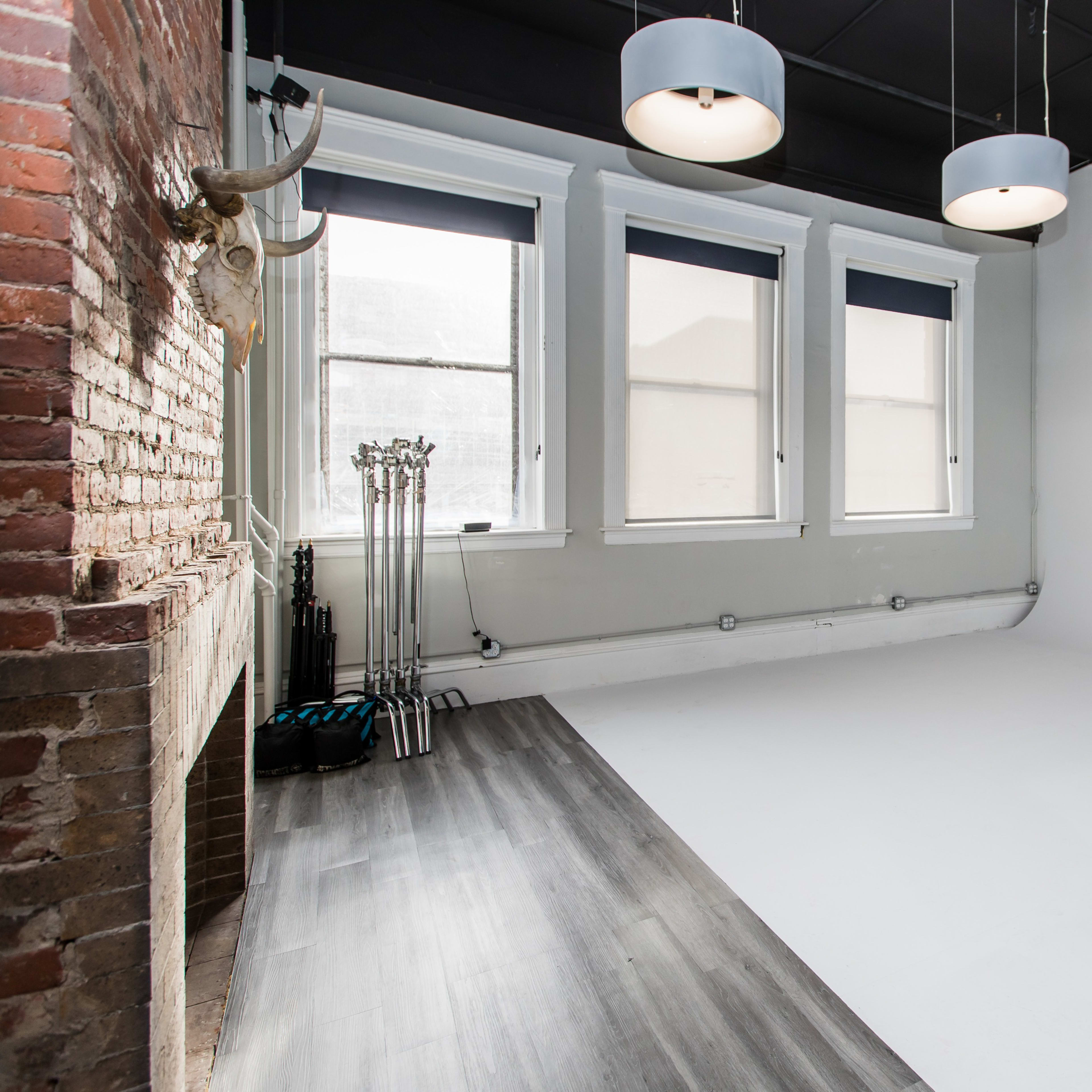 A spacious room with brick walls, large windows, and a white wall, featuring modern light fixtures and a wooden floor.