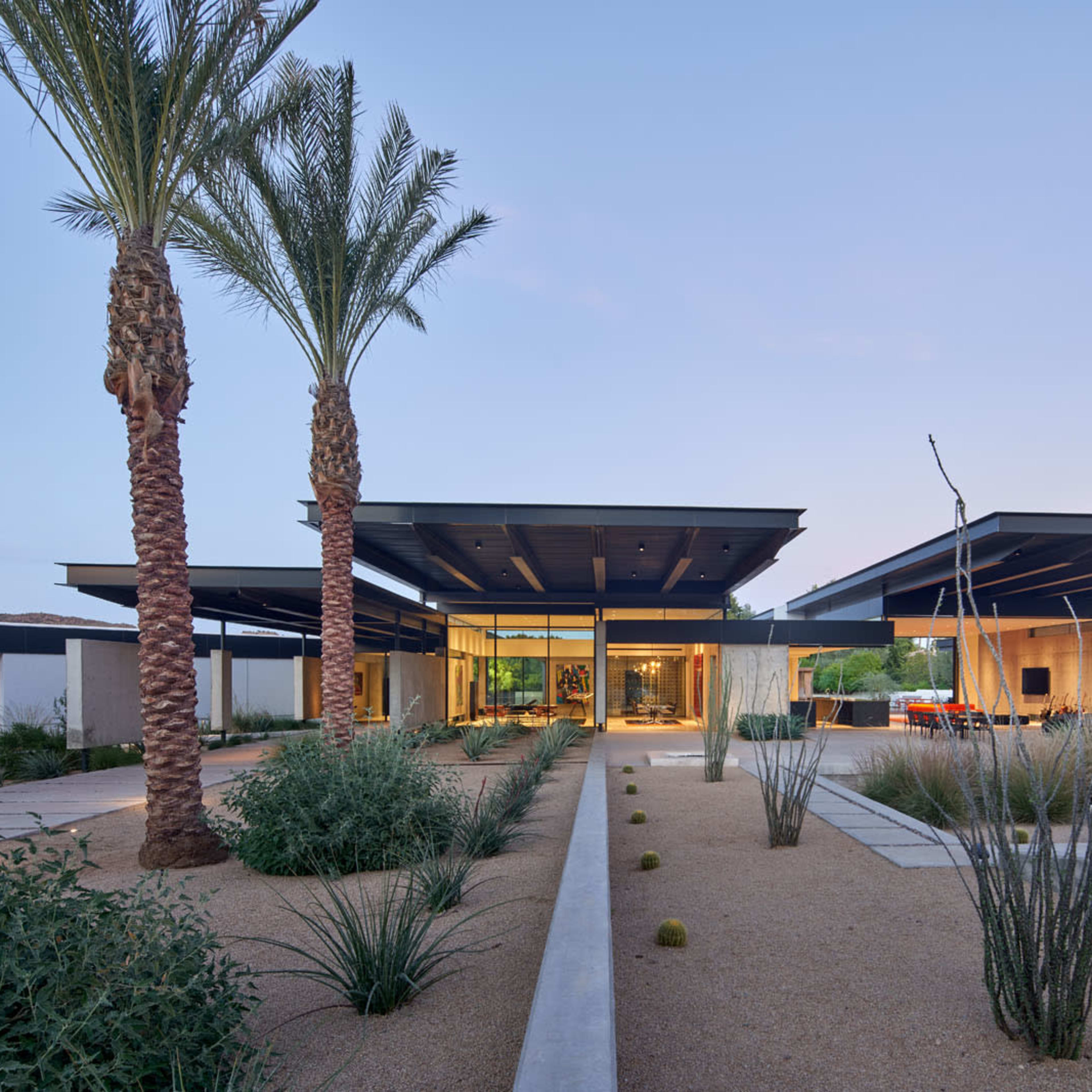 A modern, single-story house features expansive flat roofs and is surrounded by desert landscaping, including palm trees and low shrubs.