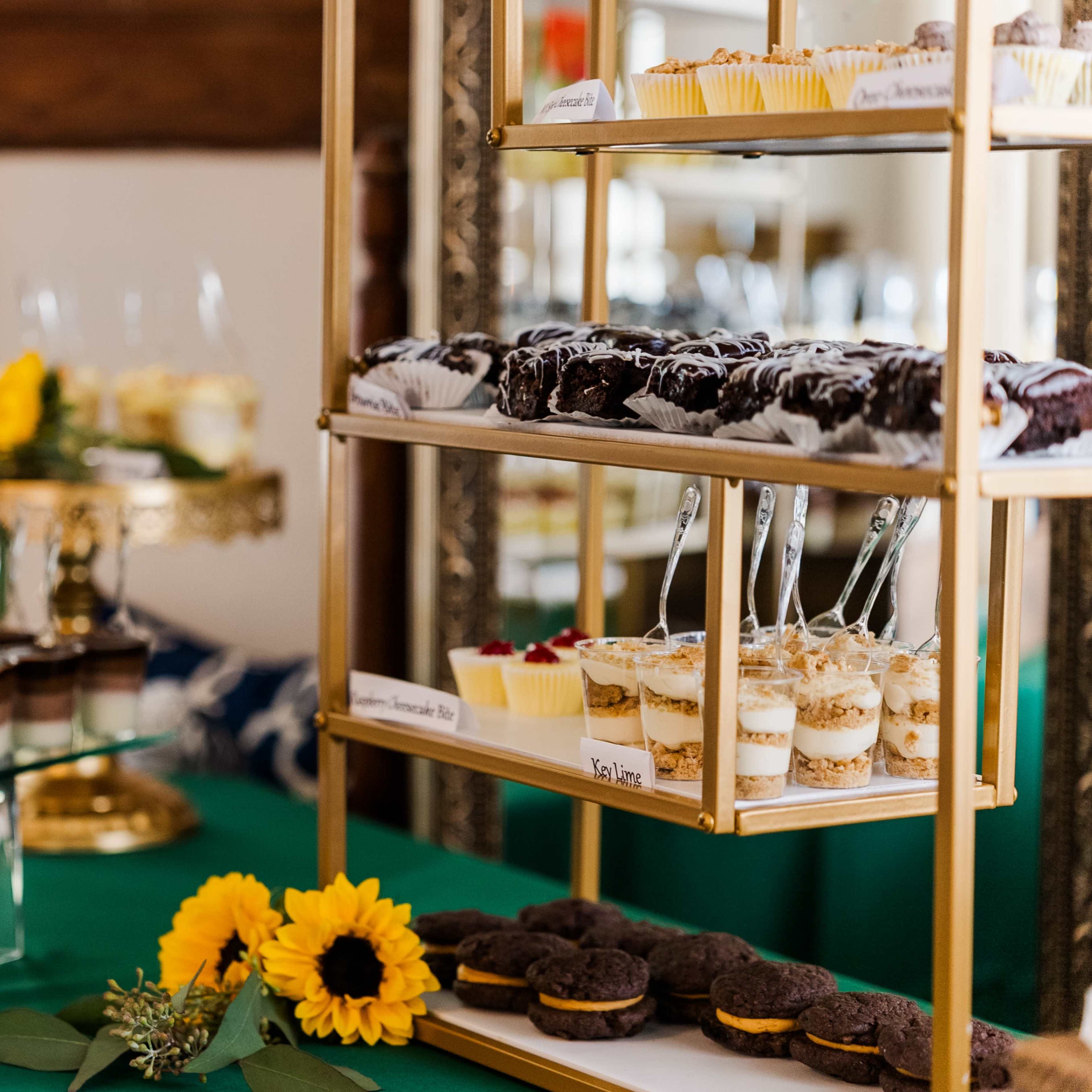 The display features a three-tiered stand filled with various desserts, including cupcakes, layered parfaits, and cookies, arranged alongside fresh flowers and greenery.