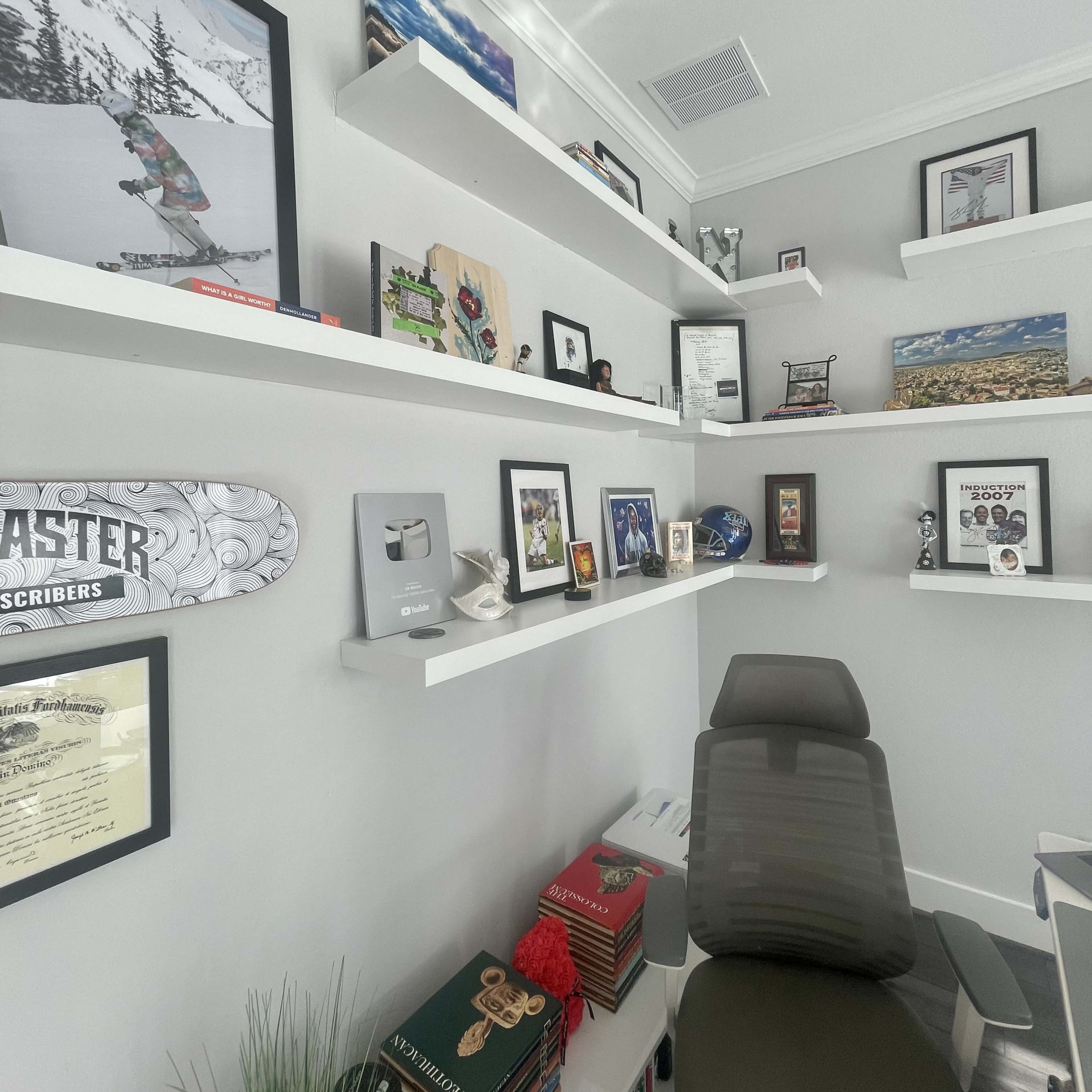 A modern office space featuring white shelves adorned with various framed pictures, awards, and decorative items, along with a chair and a small plant.