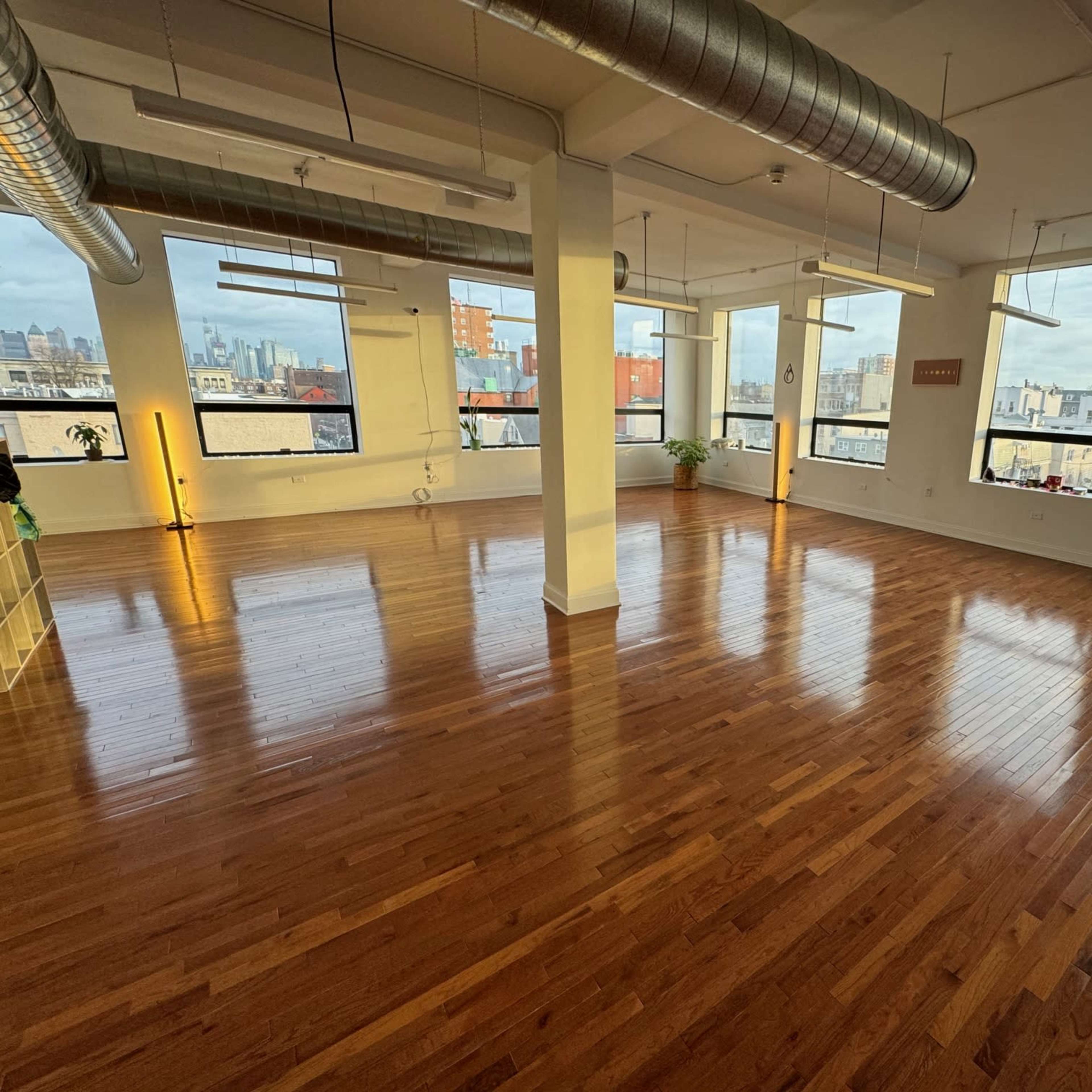 A spacious, empty room with polished wooden floors and large windows offering views of the city skyline.