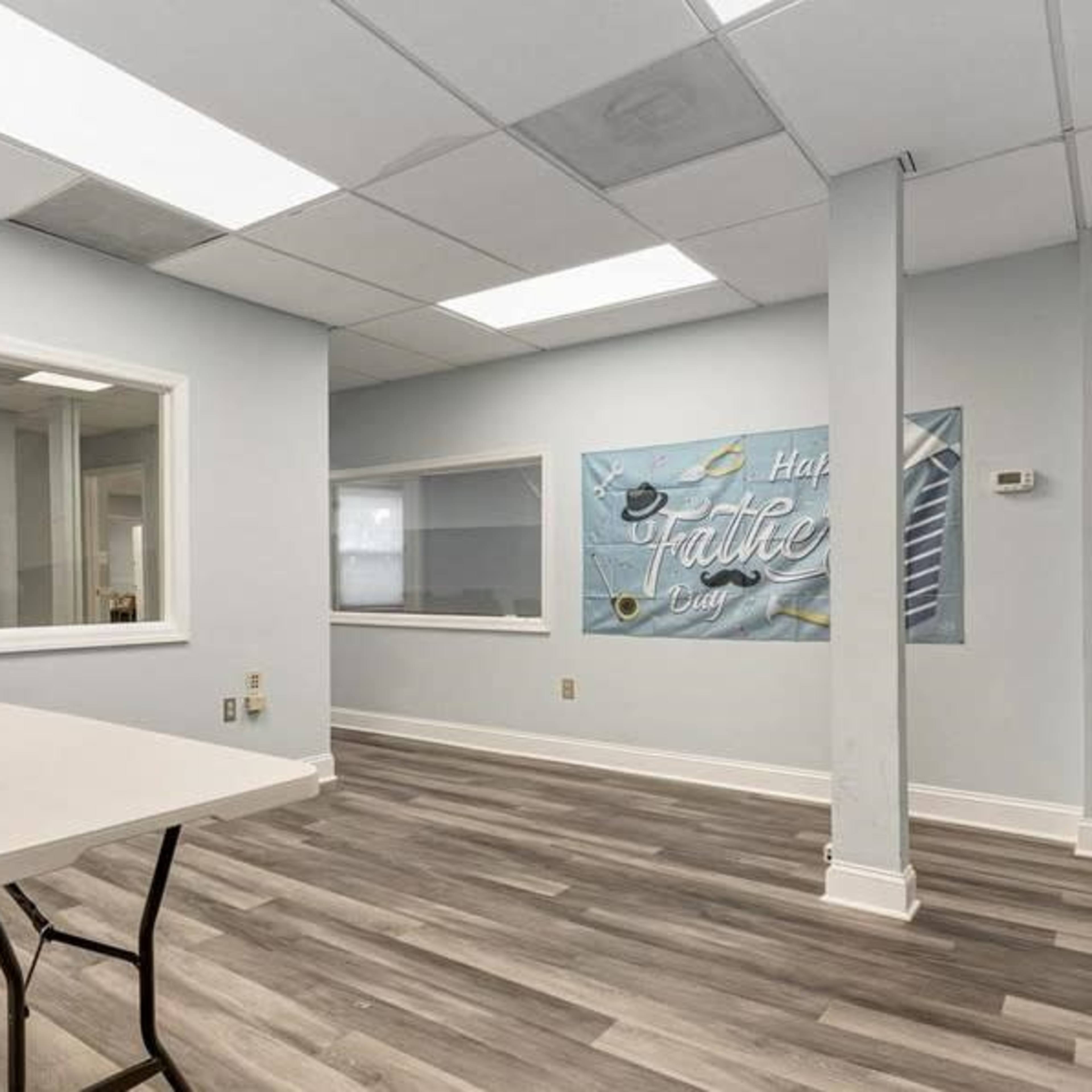 A sparsely furnished office space features a table, gray walls, and large windows with a wall mural that reads "Half Baked Cafe."