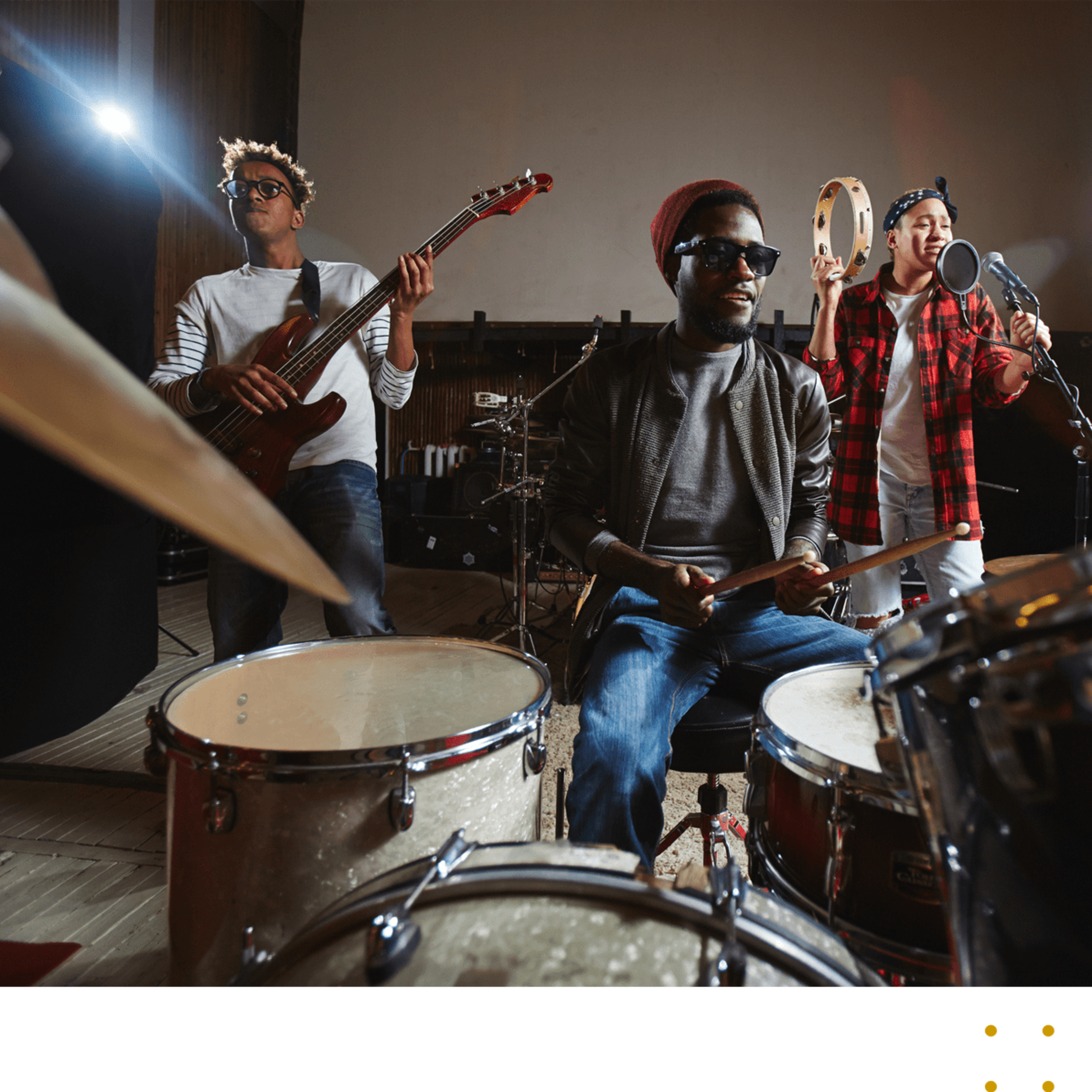 Three musicians perform together in a recording studio, with one playing the drums, another on bass, and a third singing into a microphone.