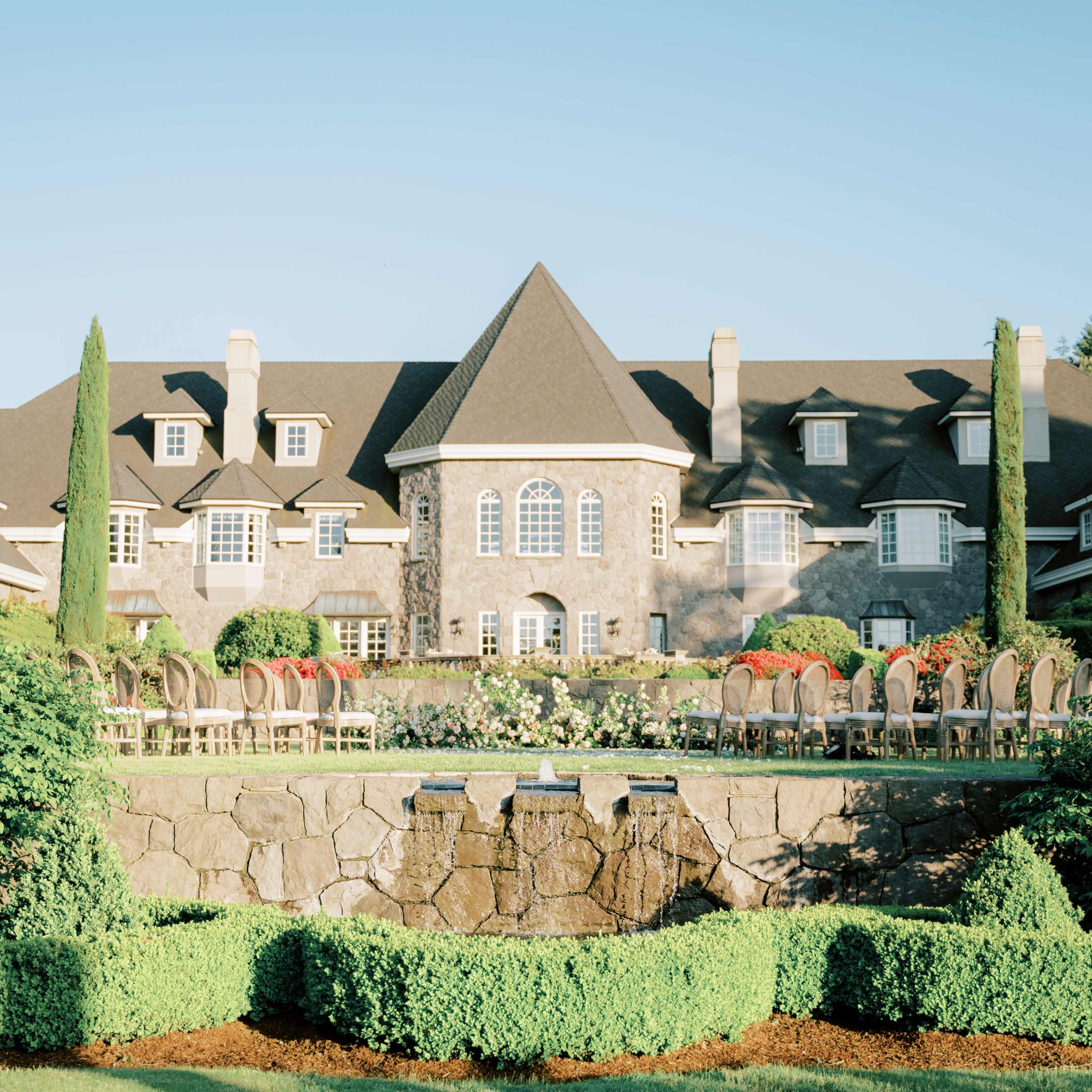 A large stone mansion with a steeply pitched roof, surrounded by well-maintained gardens and seating arranged on a terrace.