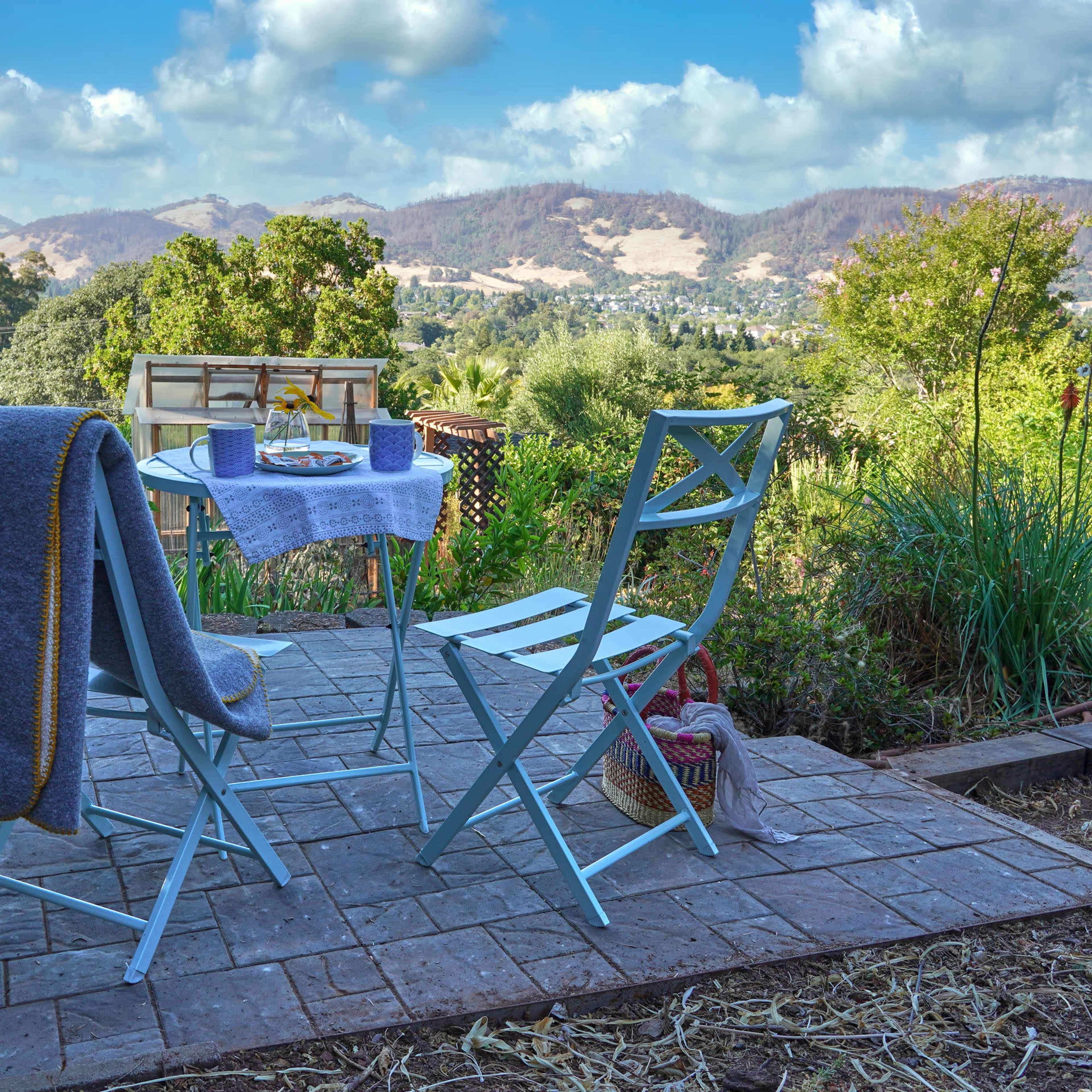 A small patio area features two light blue chairs and a table set for a meal, overlooking a lush landscape and distant mountains under a partly cloudy sky.