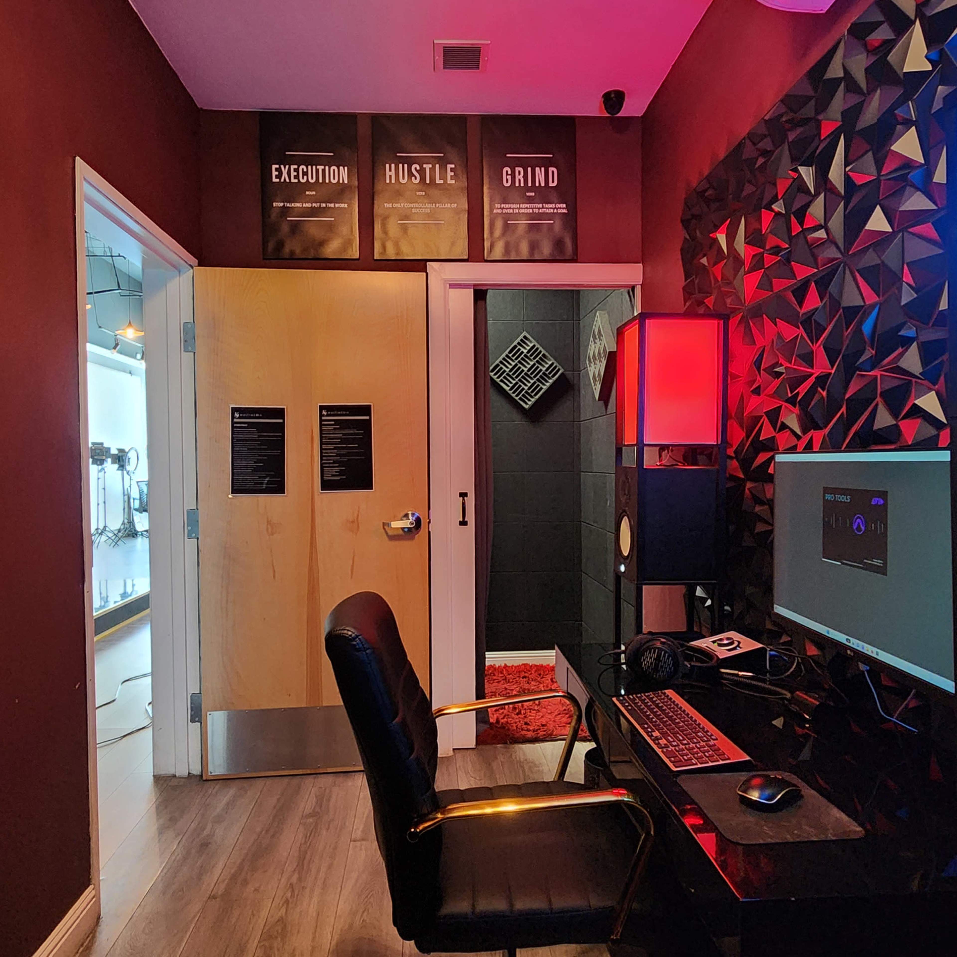 A recording studio with a desk, computer monitor, and speakers, featuring a wall with geometric patterns and motivational signs above the door.