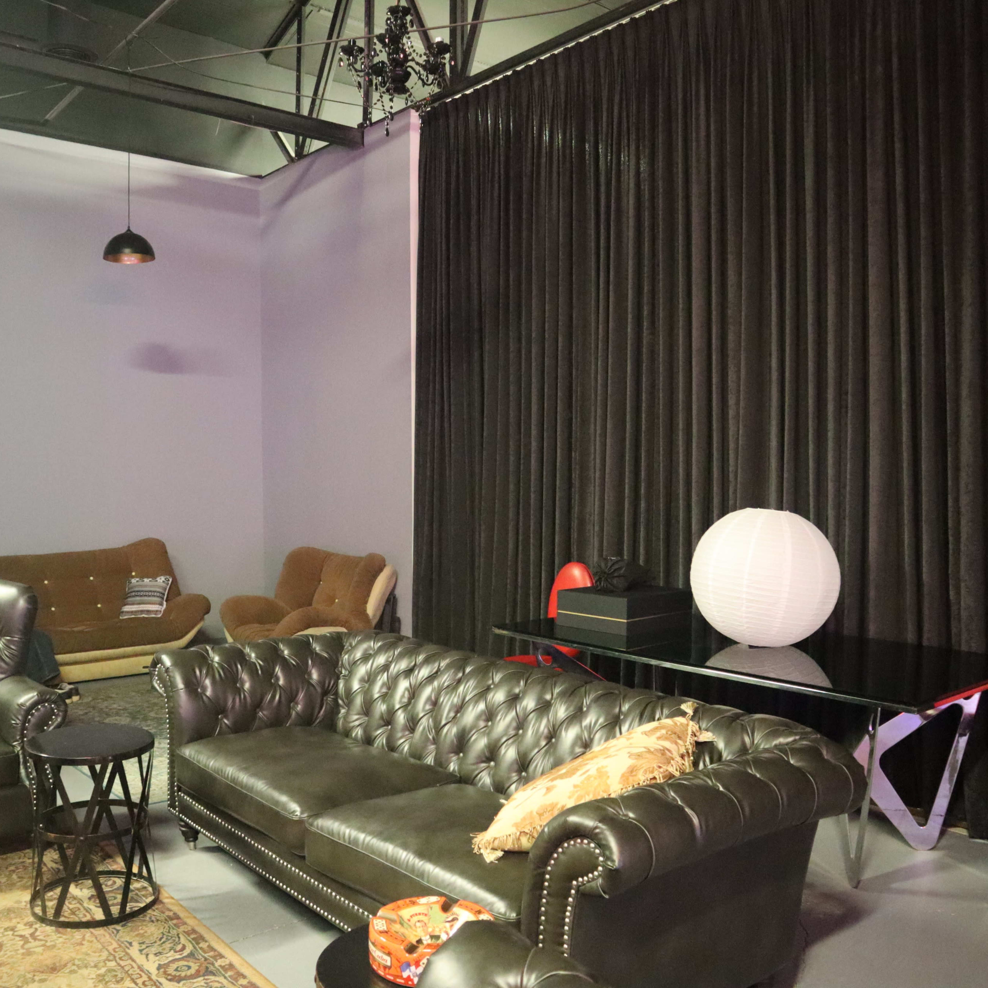 A modern lounge area with a black leather couch, two upholstered chairs, a round side table, and a white paper lantern on a black table, all set against a backdrop of black curtains.