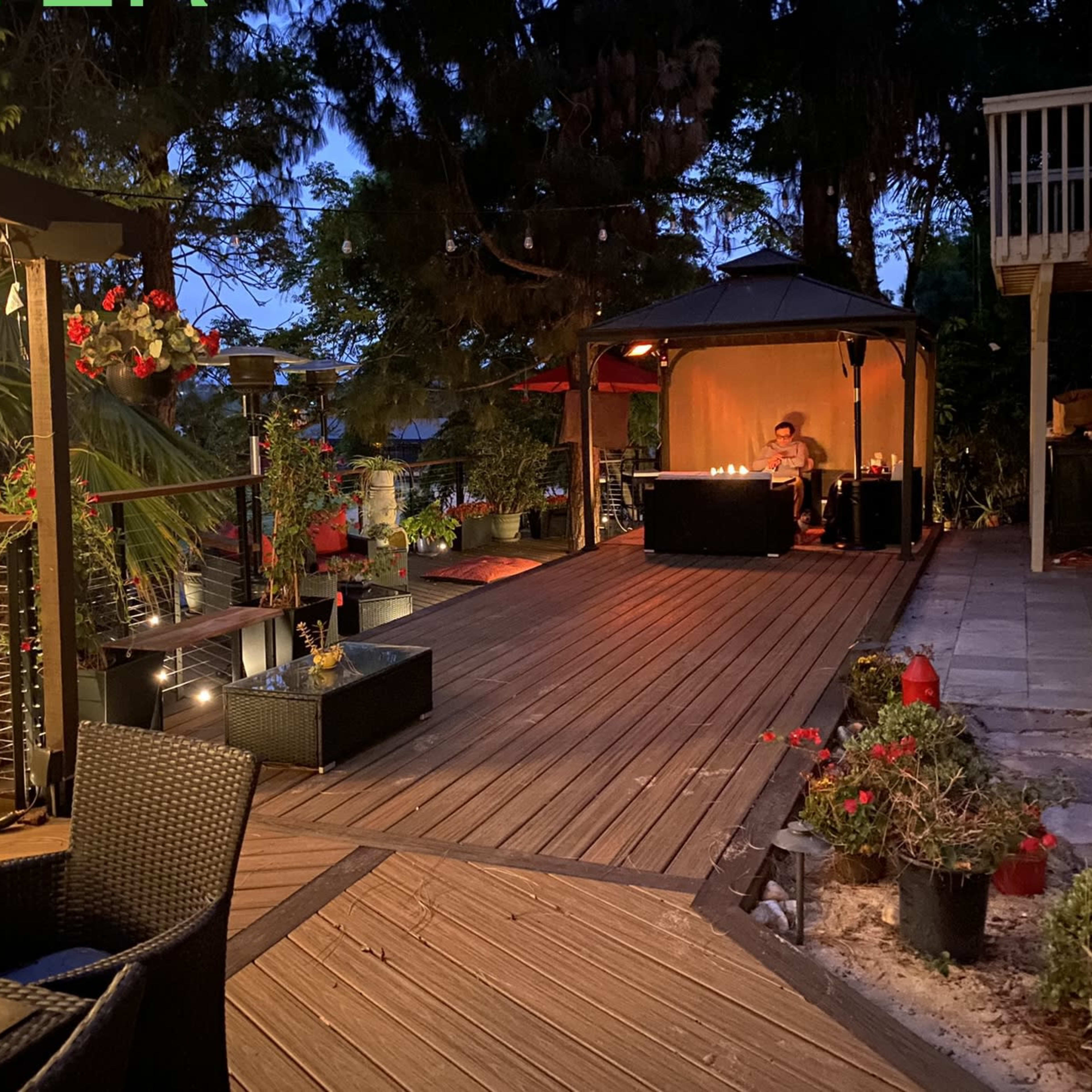 A wooden deck at dusk features a gazebo with a table set for dinner and potted plants along the walkway.