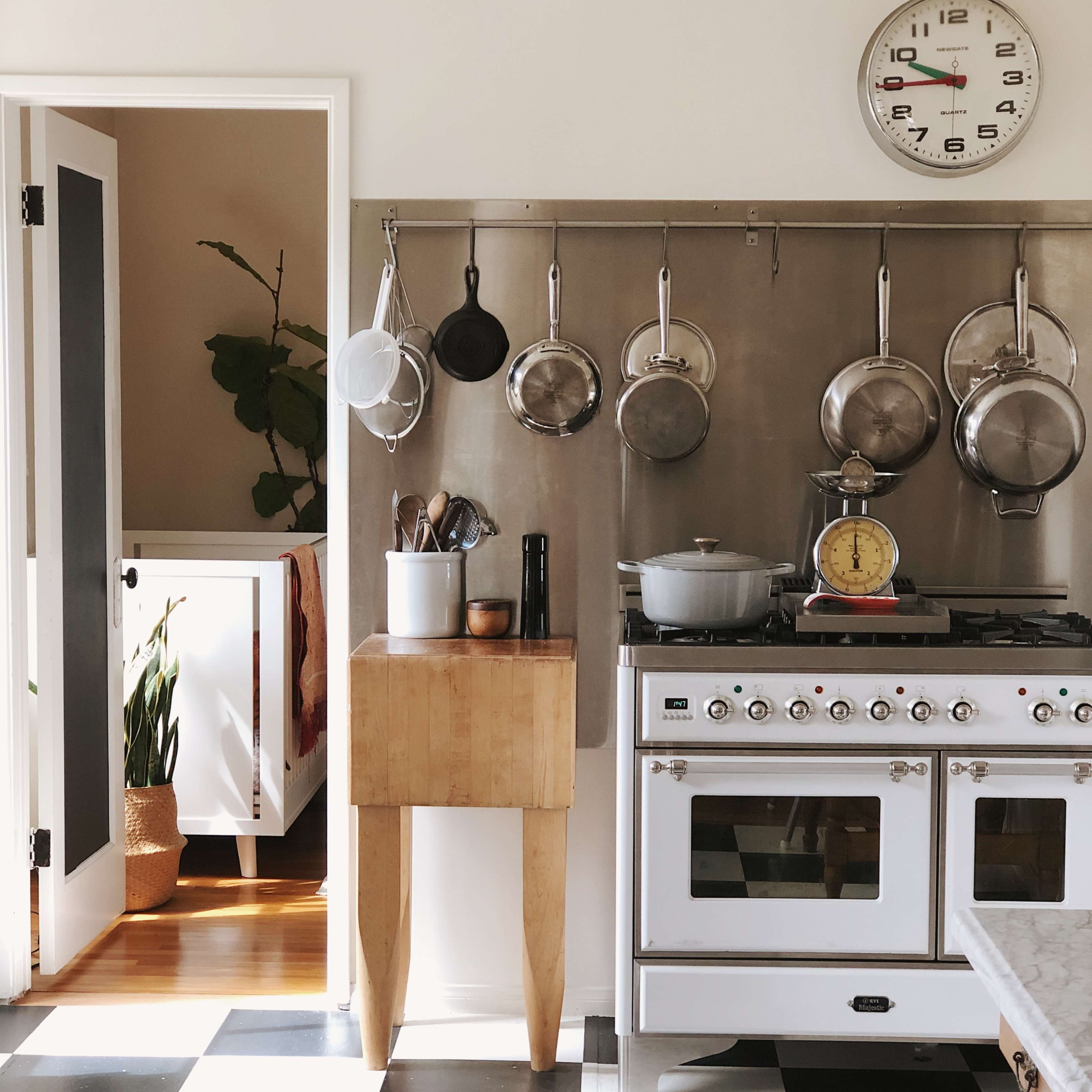 A kitchen with a stainless steel stove, hanging frying pans on a wall, and a wooden table beside a doorway leading to another room.