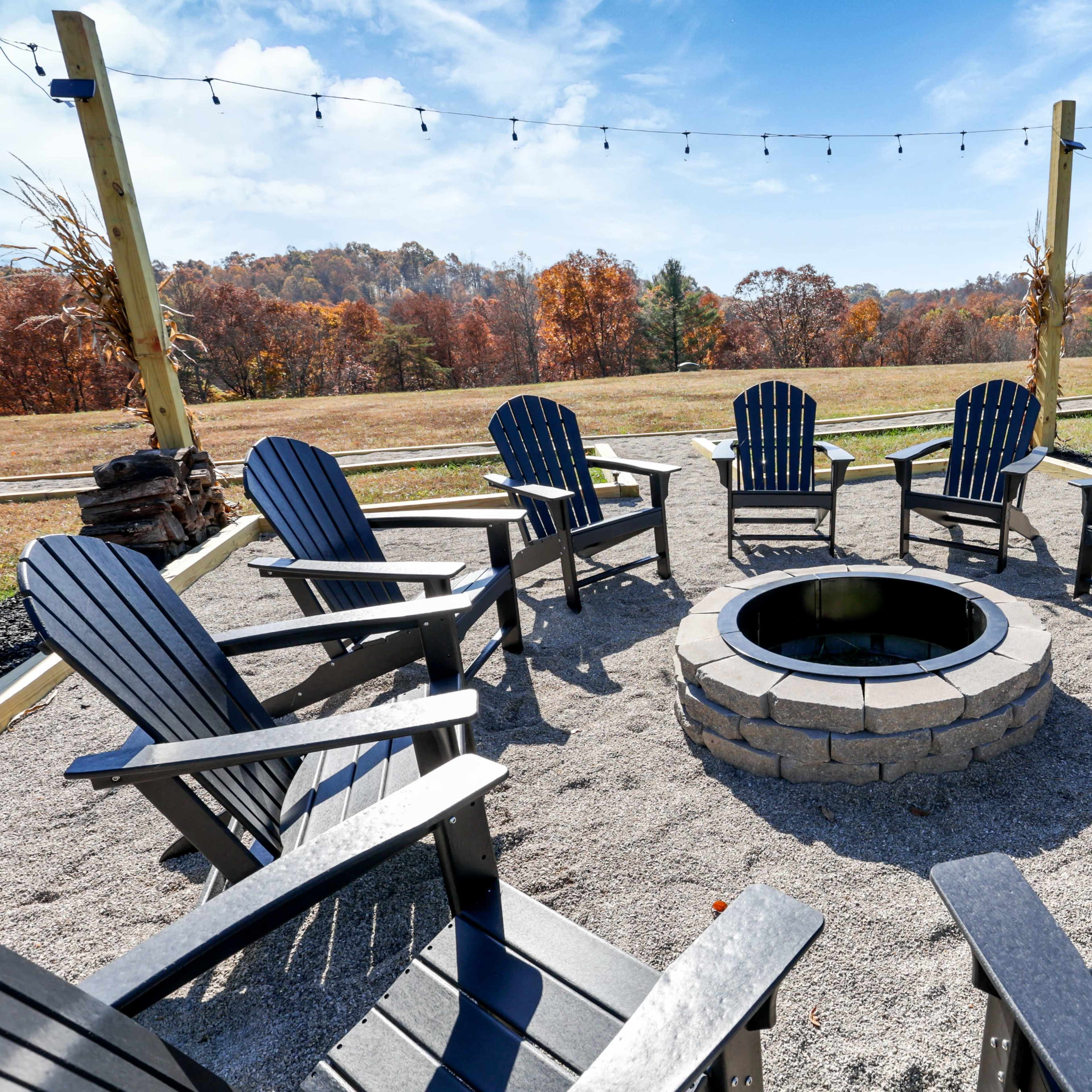 A circular seating area with black Adirondack chairs surrounds a stone fire pit, set on gravel with a view of autumn foliage in the background.