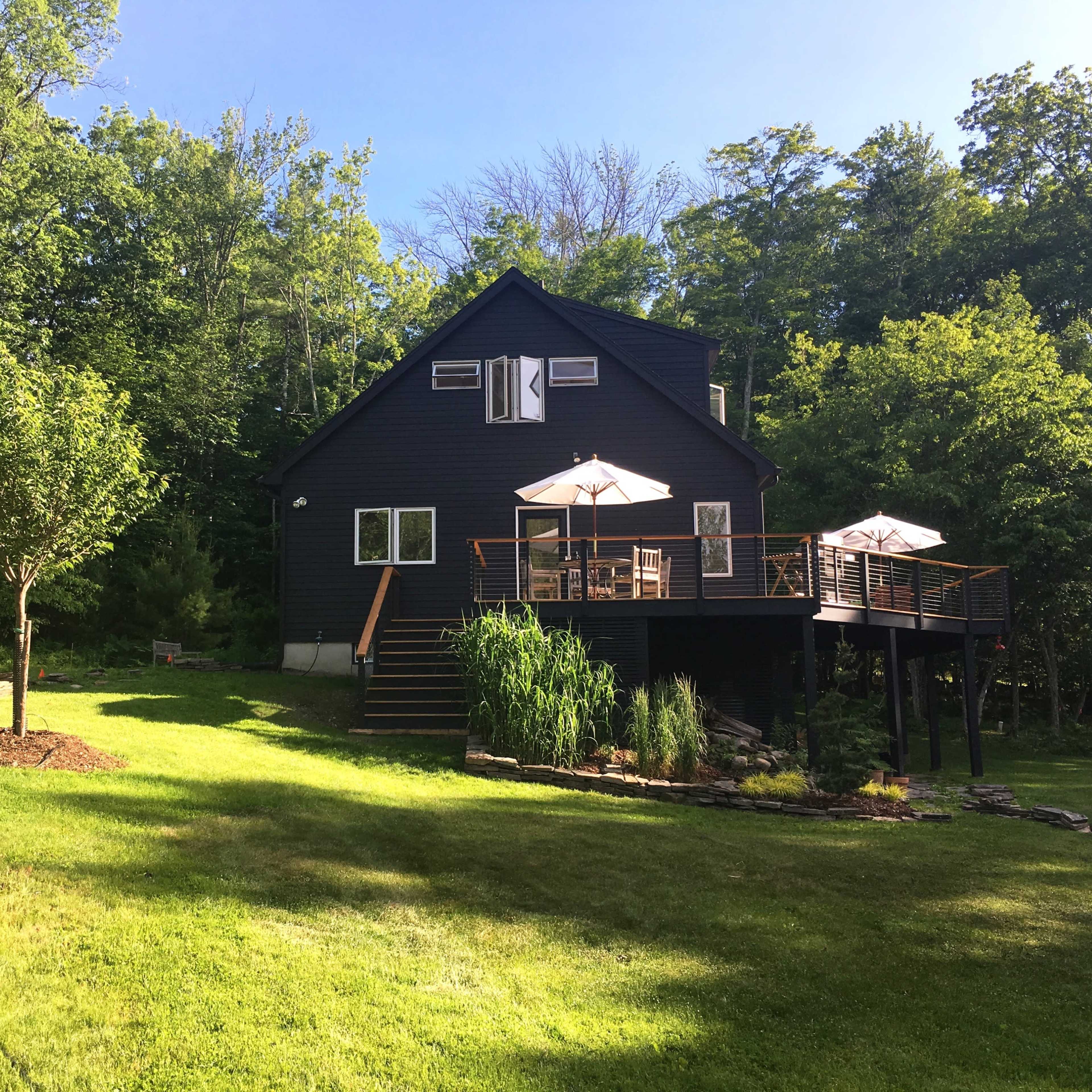 A black wooden house with a deck and patio umbrellas stands in a lush green yard surrounded by trees.