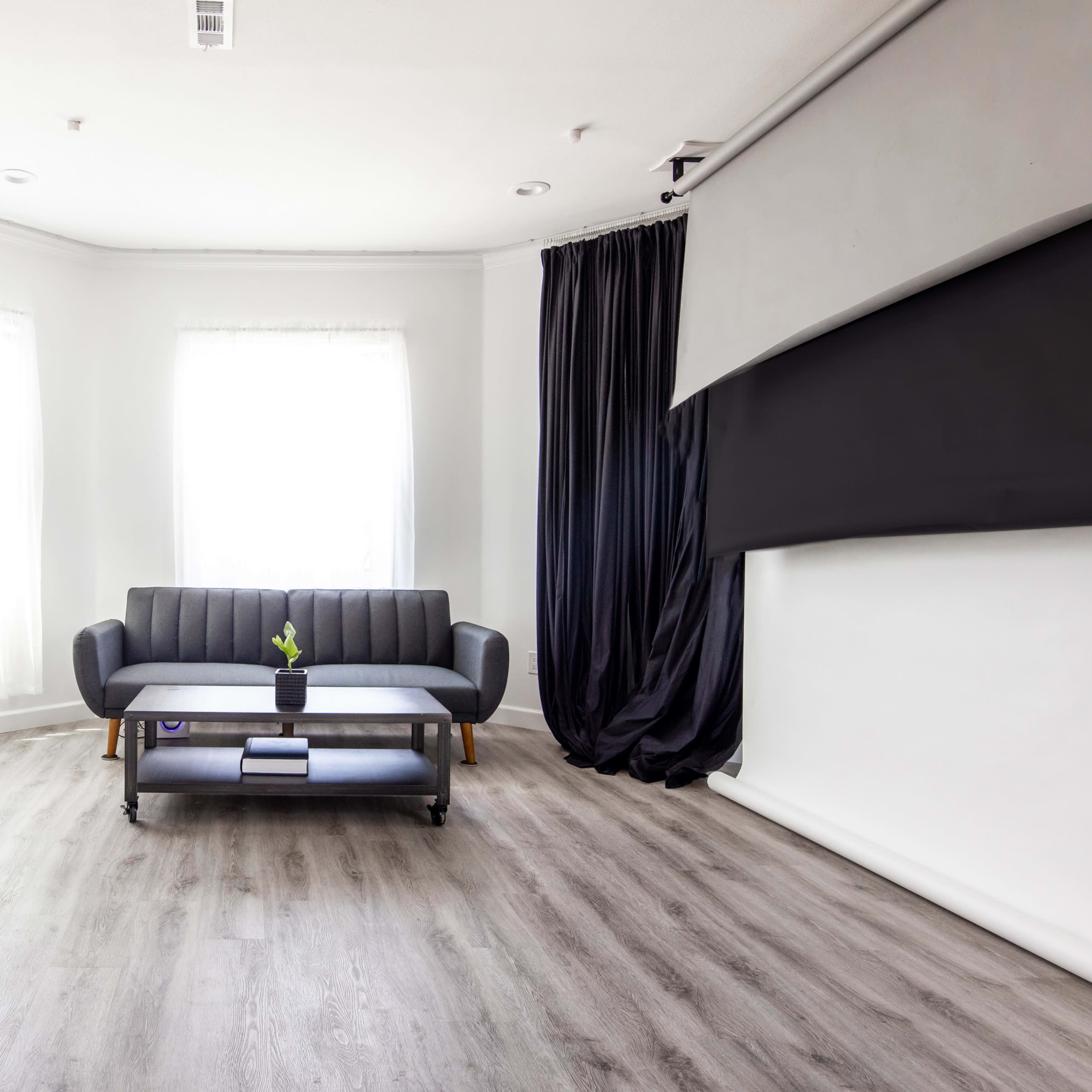 A minimalist living room featuring a gray sofa, a small coffee table with a potted plant, large windows with white curtains, and a dark curtain covering part of a wall.