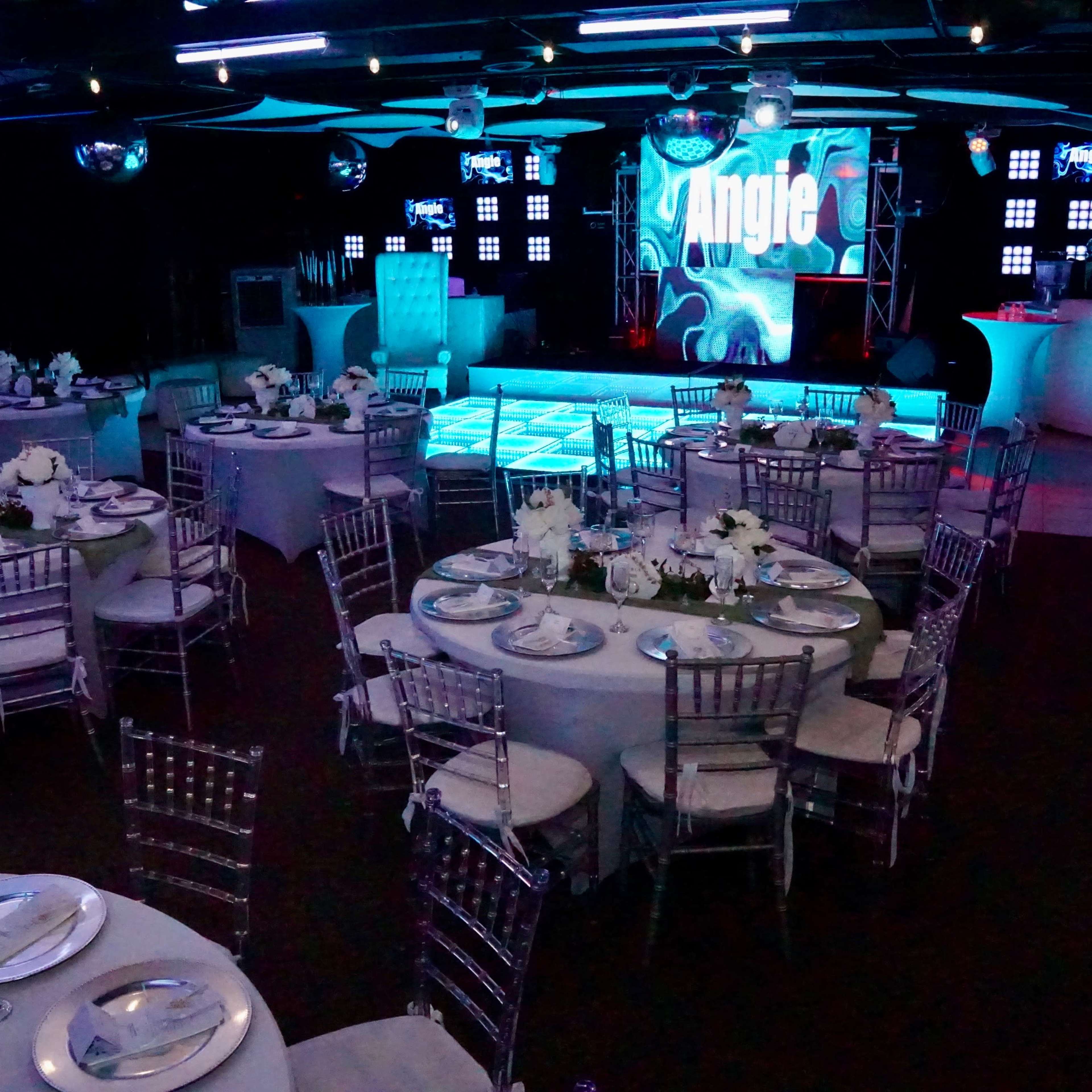 A banquet hall set up for an event, featuring circular tables with white tablecloths and floral centerpieces, a lit dance floor, and a large screen displaying the name "Angie."