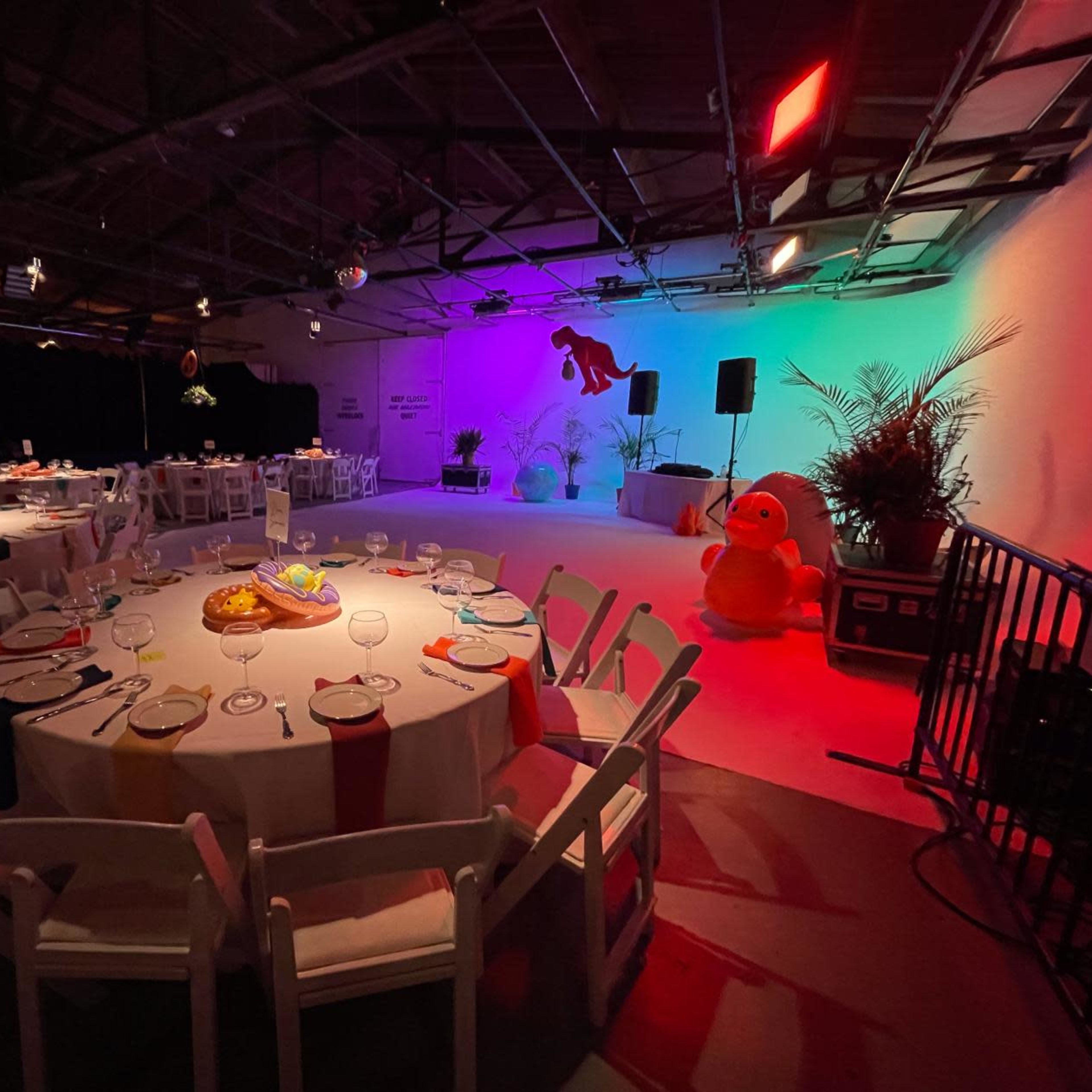 A decorated event space with round tables set for dining, colorful lighting, and stage elements in the background.
