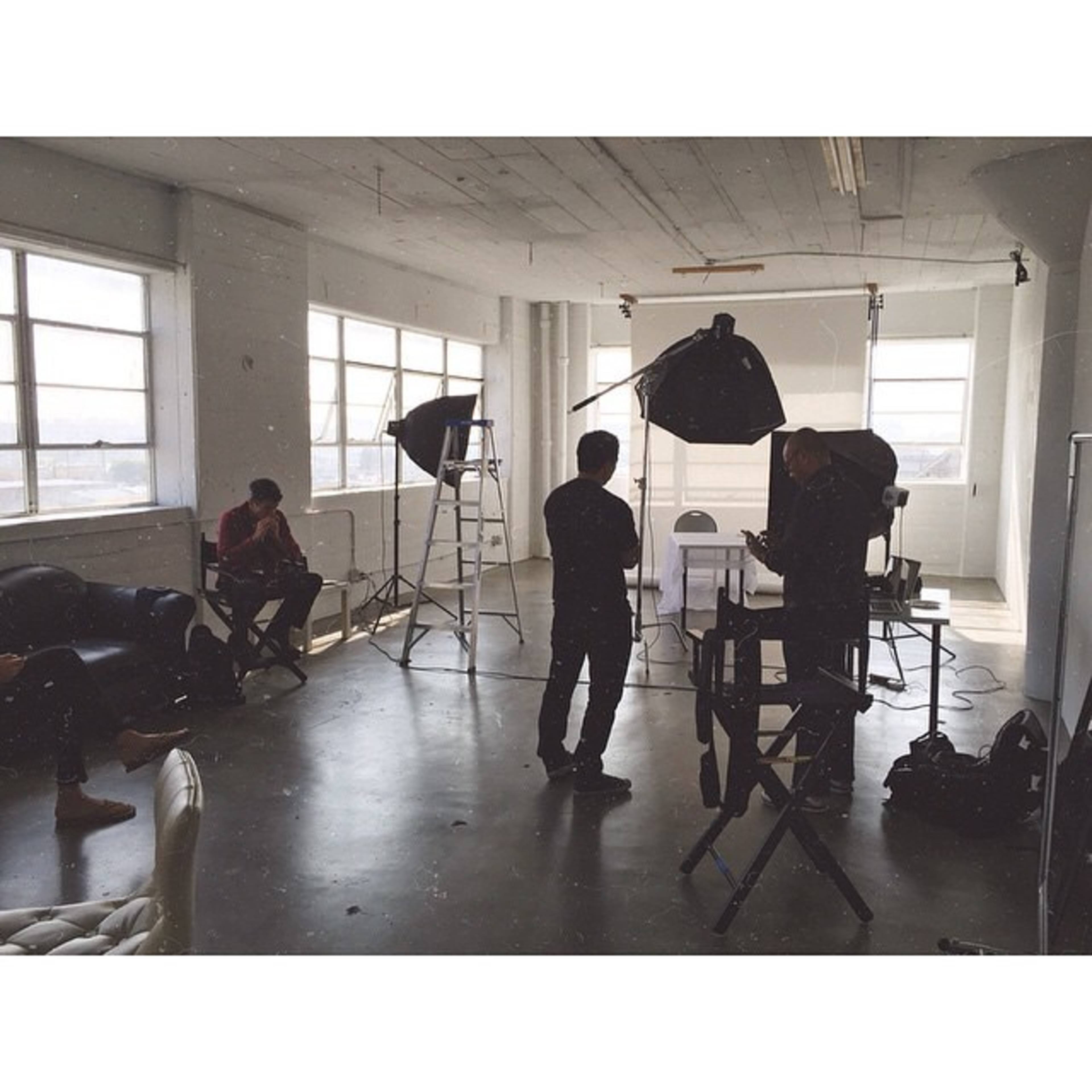 A photography studio setting with individuals preparing for a photoshoot, featuring lighting equipment and a backdrop.