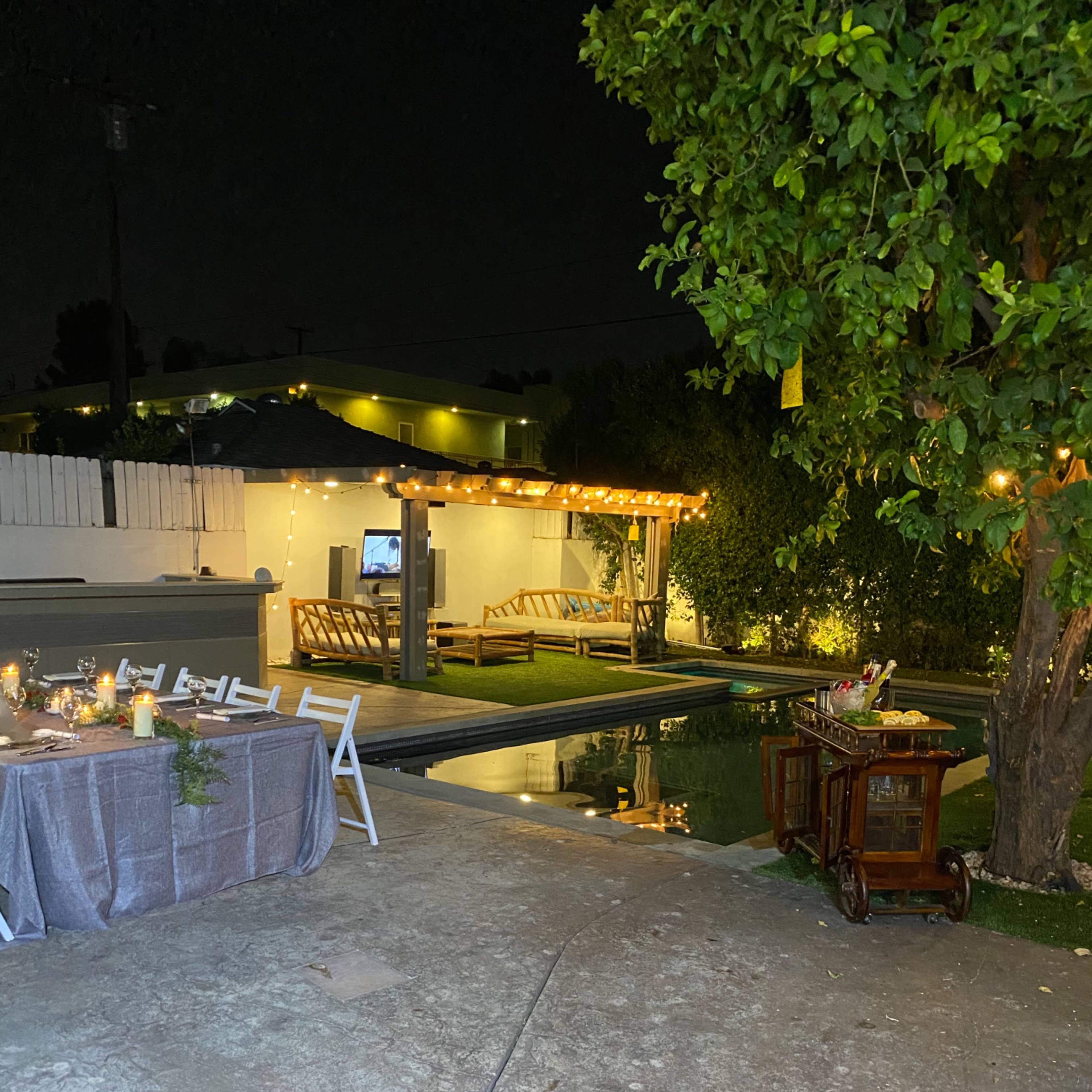 A dimly lit outdoor space features a dining table, a pool, and a wooden pergola with string lights.